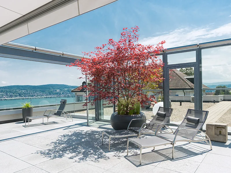Paved roof terrace with lounge chairs and a red flowering tree overlooking Lake Zurich