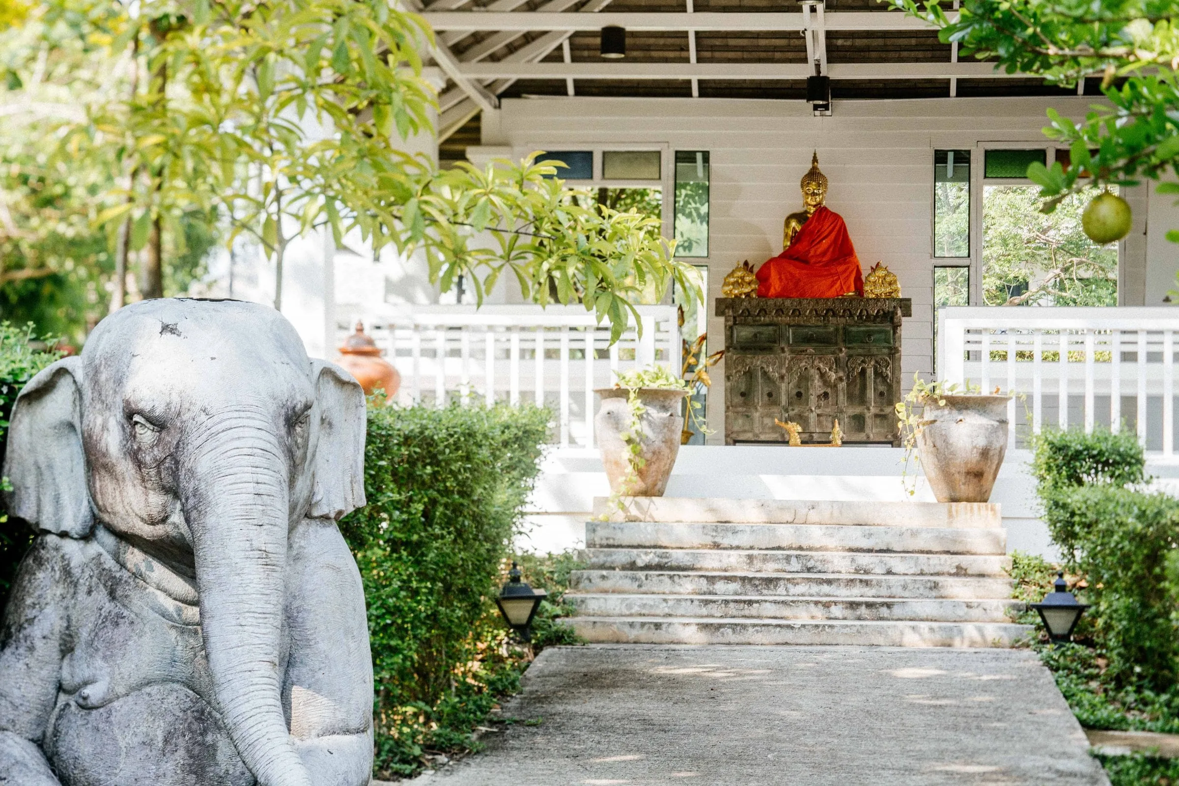 Stone elephant statue beside garden steps leading up to a white veranda with a seated Buddha shrine