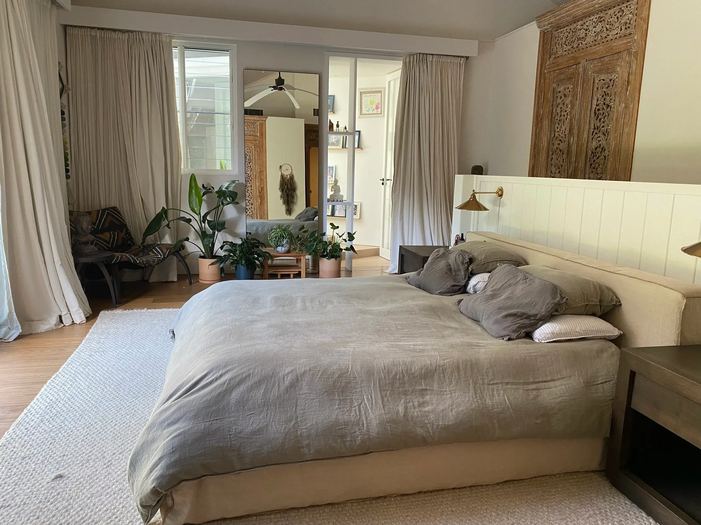 Master bedroom with a grey linen bed, carved timber wall panel, sheer curtains and potted plants