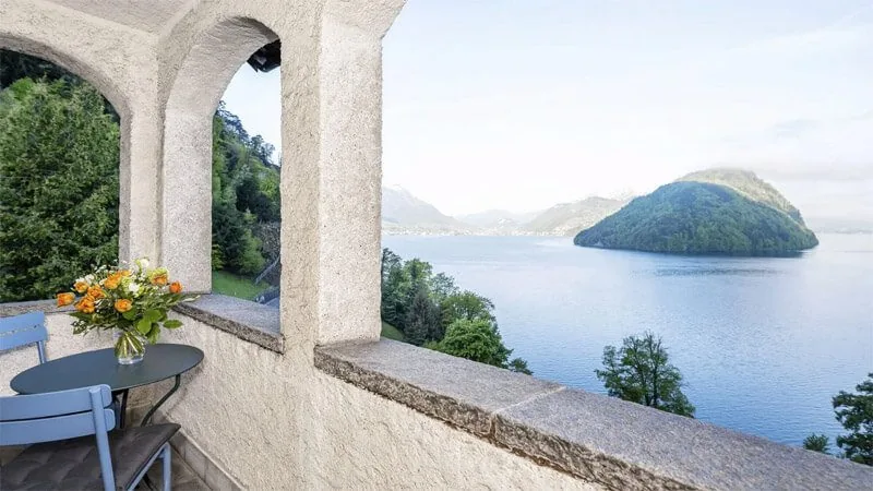 Stone arched balcony with a small table and flowers looking out over Lake Lucerne and a wooded island