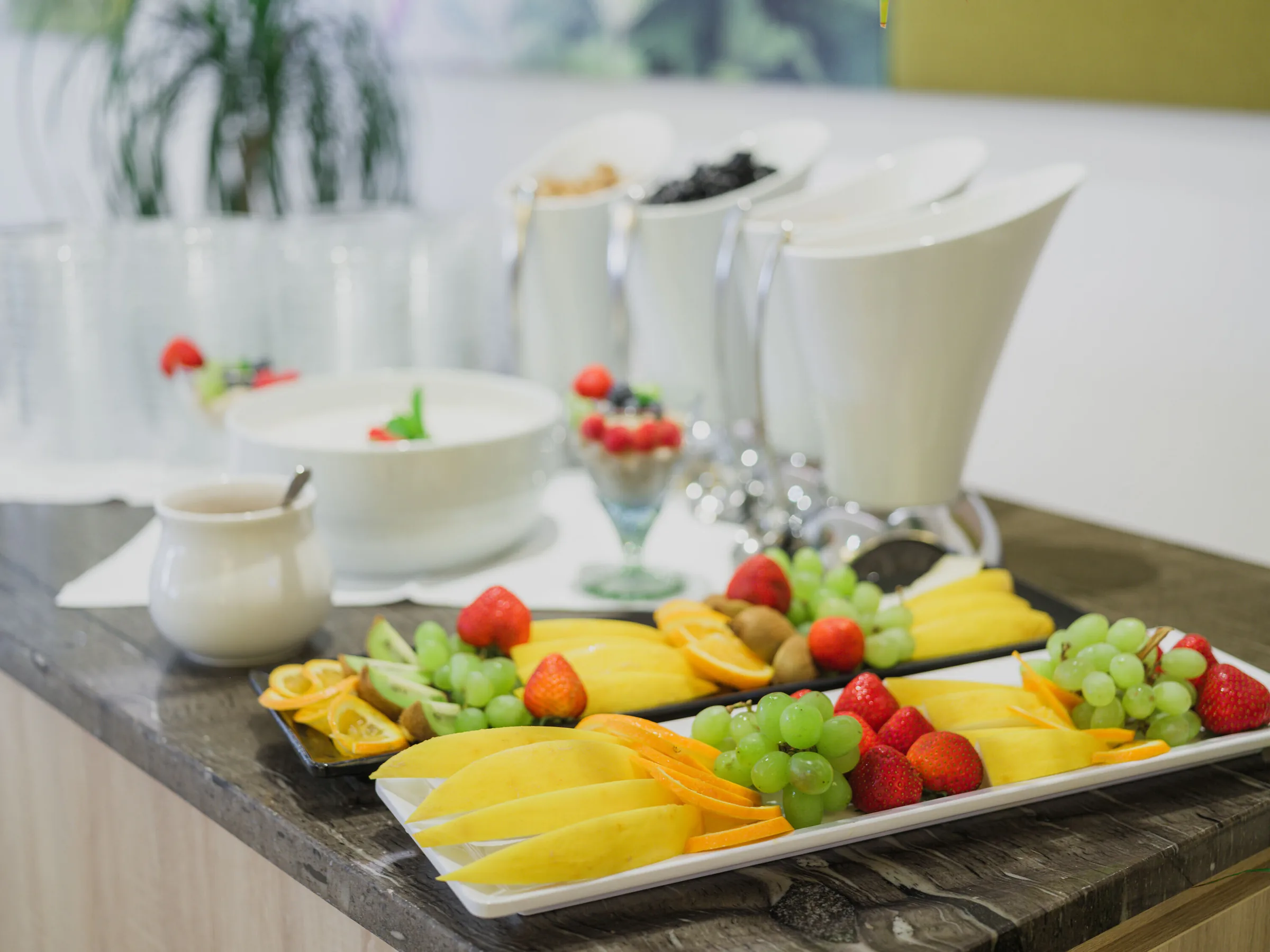 Platters of sliced mango, kiwi, grapes and strawberries on a marble counter beside yoghurt cups