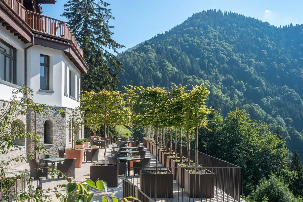Restaurant terrace with tables beside the stone building, small trees and forested hills behind