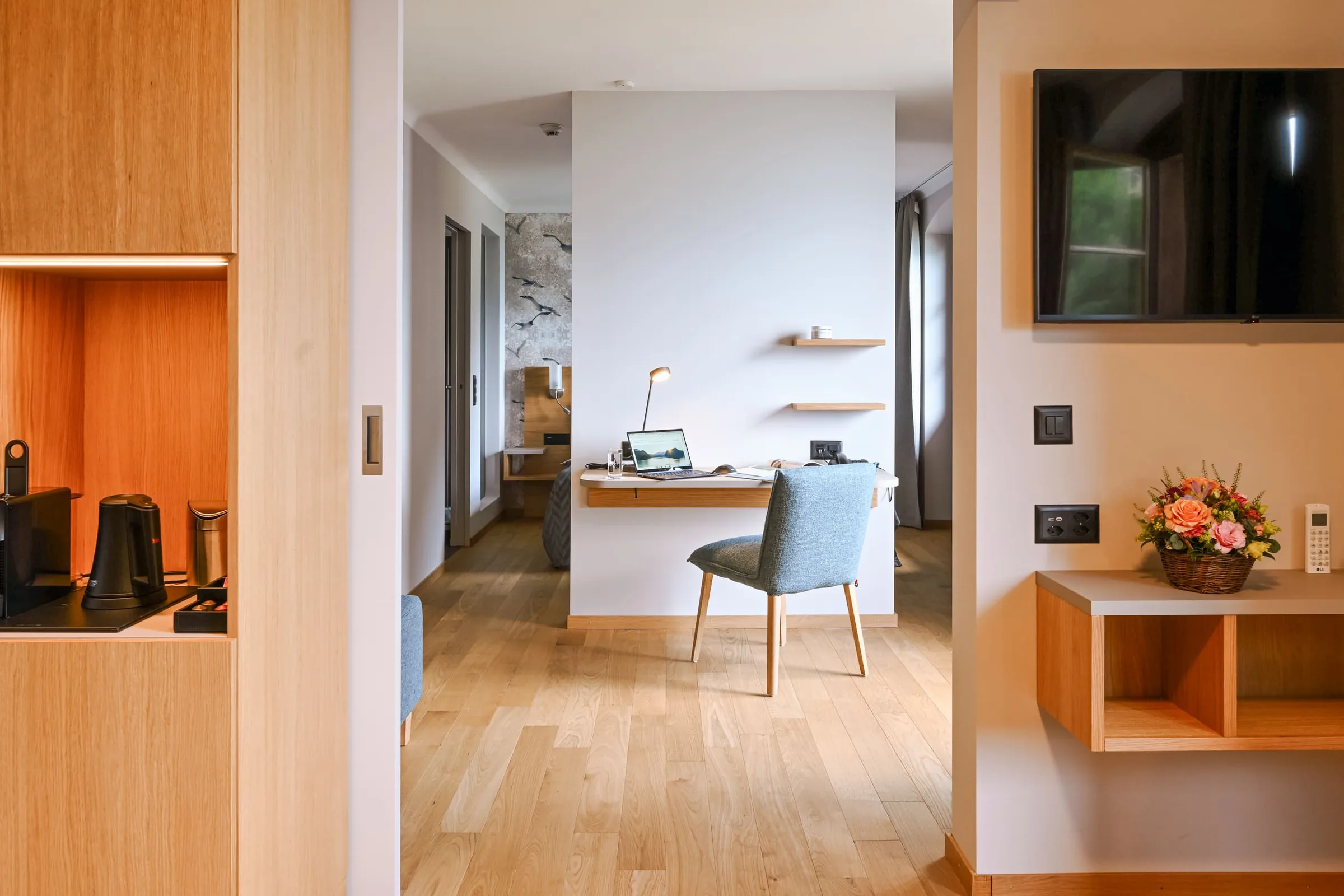 Guest room with light oak shelving, a wall mounted TV, a small desk with a blue chair and wooden floors
