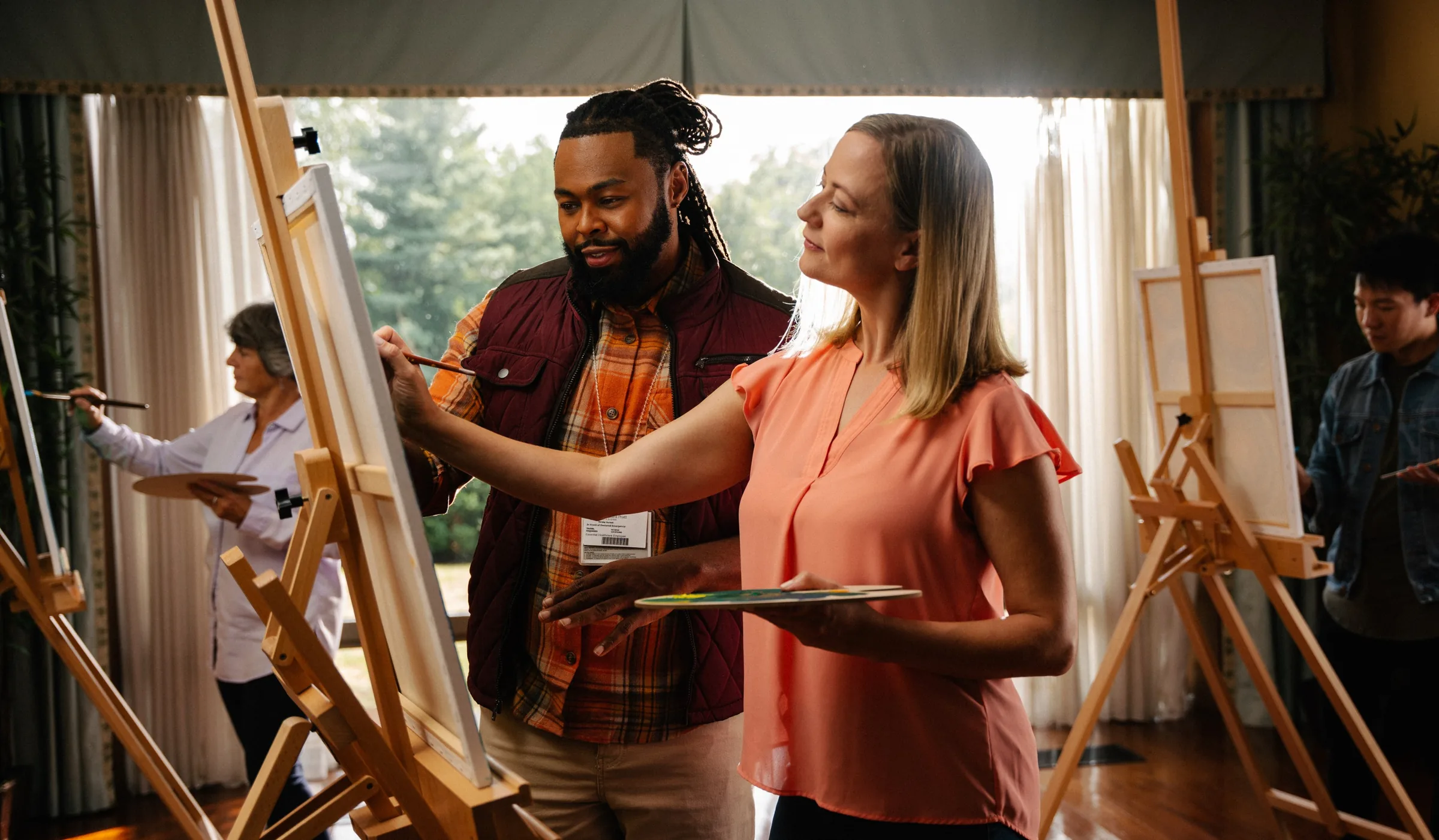 Art therapy session with several people painting at wooden easels in a room with large curtained windows