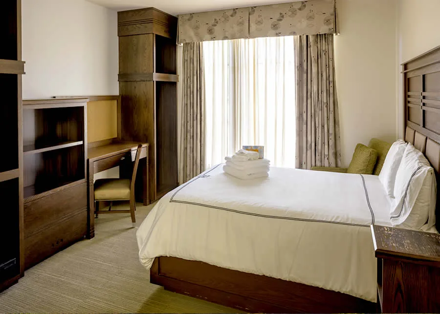 Patient room with a made bed, wooden wardrobe and desk, and a curtained window