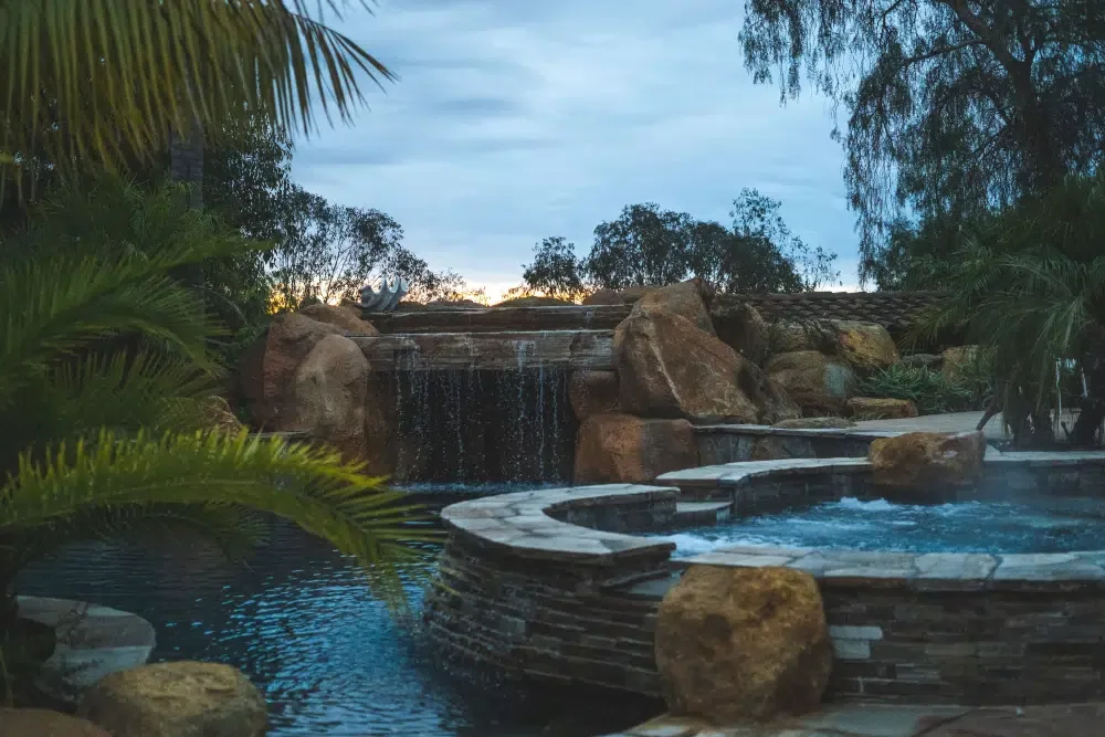 Stone-edged hot tub and pool with a rock waterfall framed by palm fronds at dusk