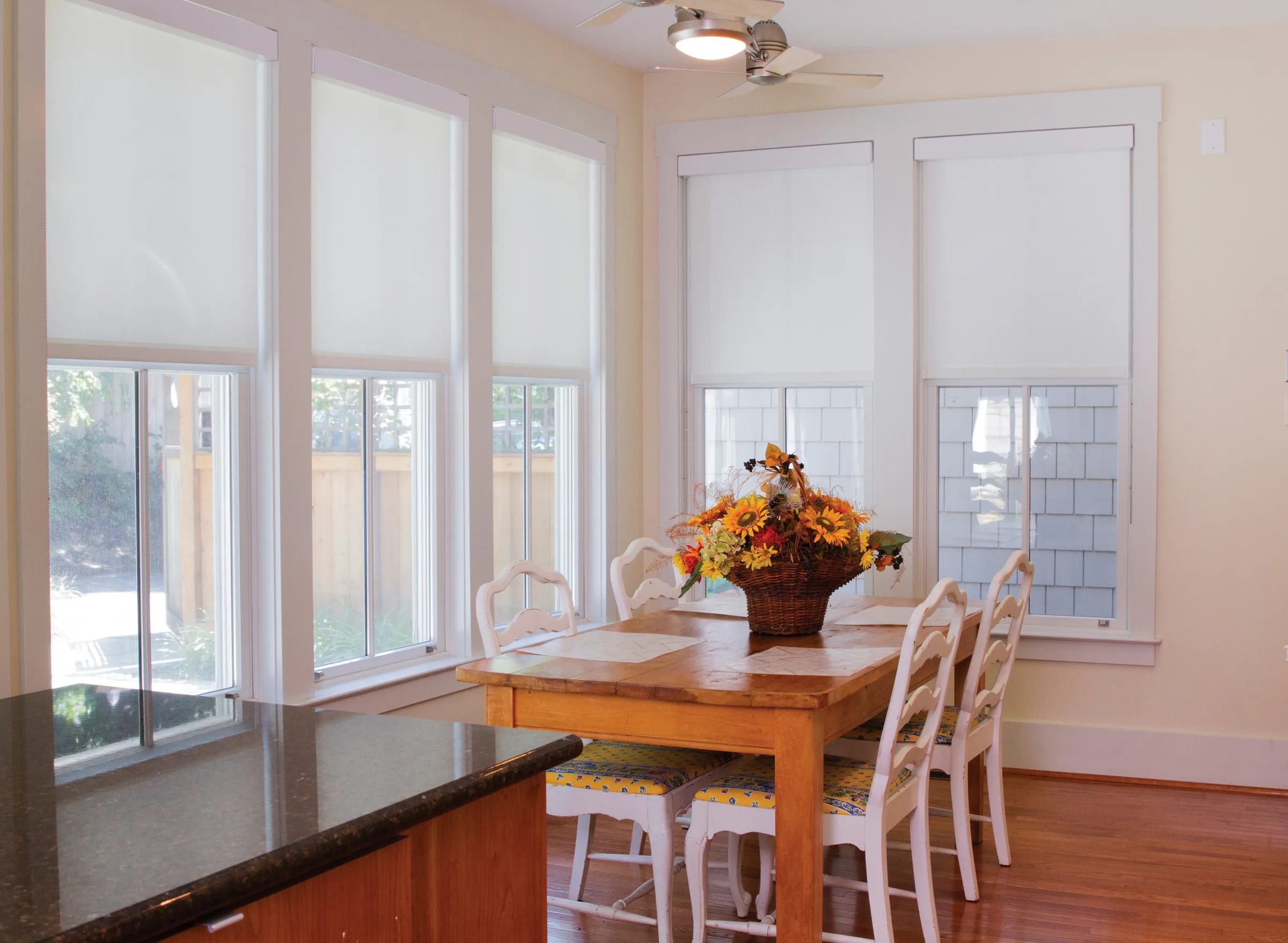 Bright dining nook with a wooden table, white chairs and flowers below three tall shaded windows