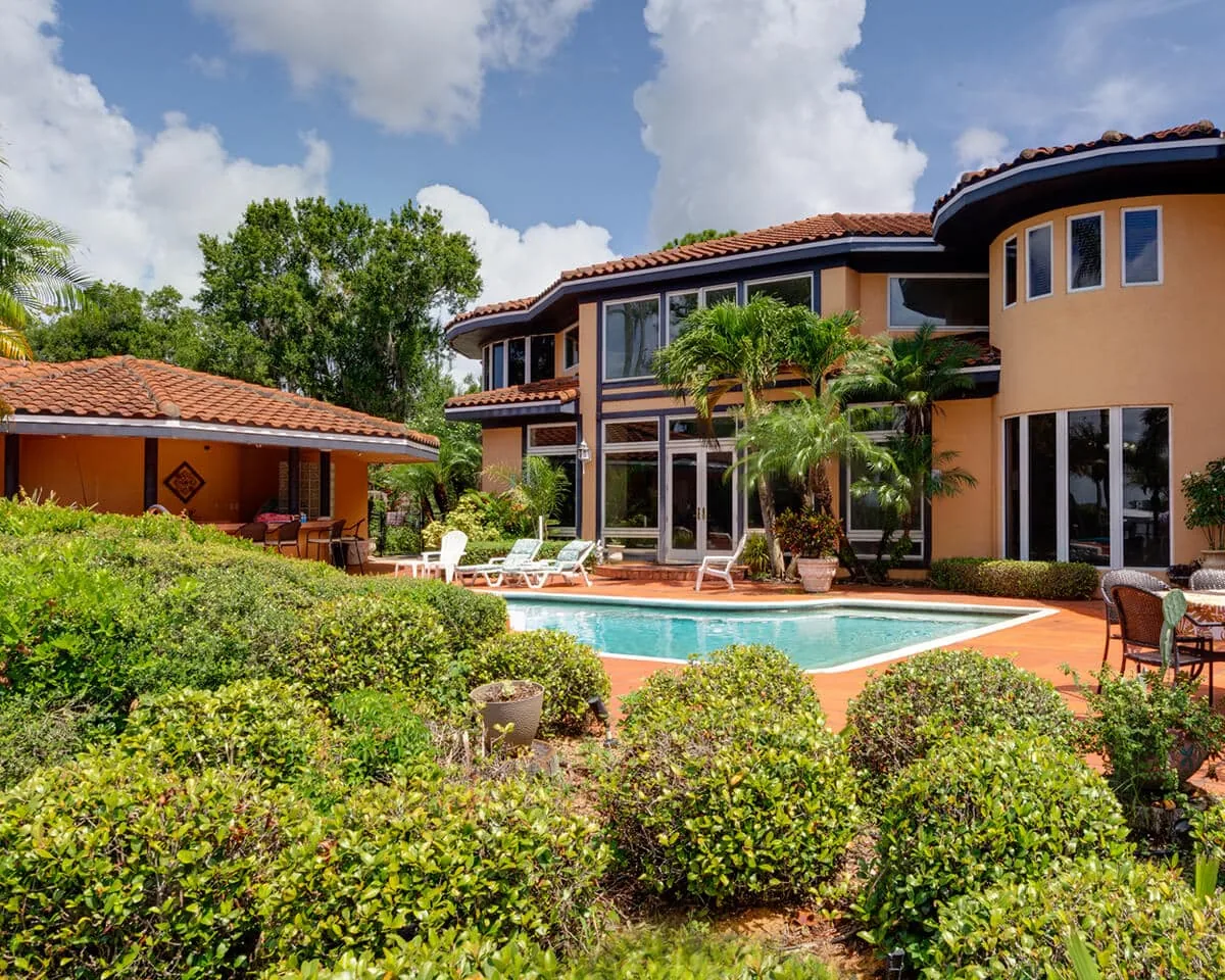 Outdoor pool with loungers behind the two storey stucco house, framed by flowering shrubs and palms