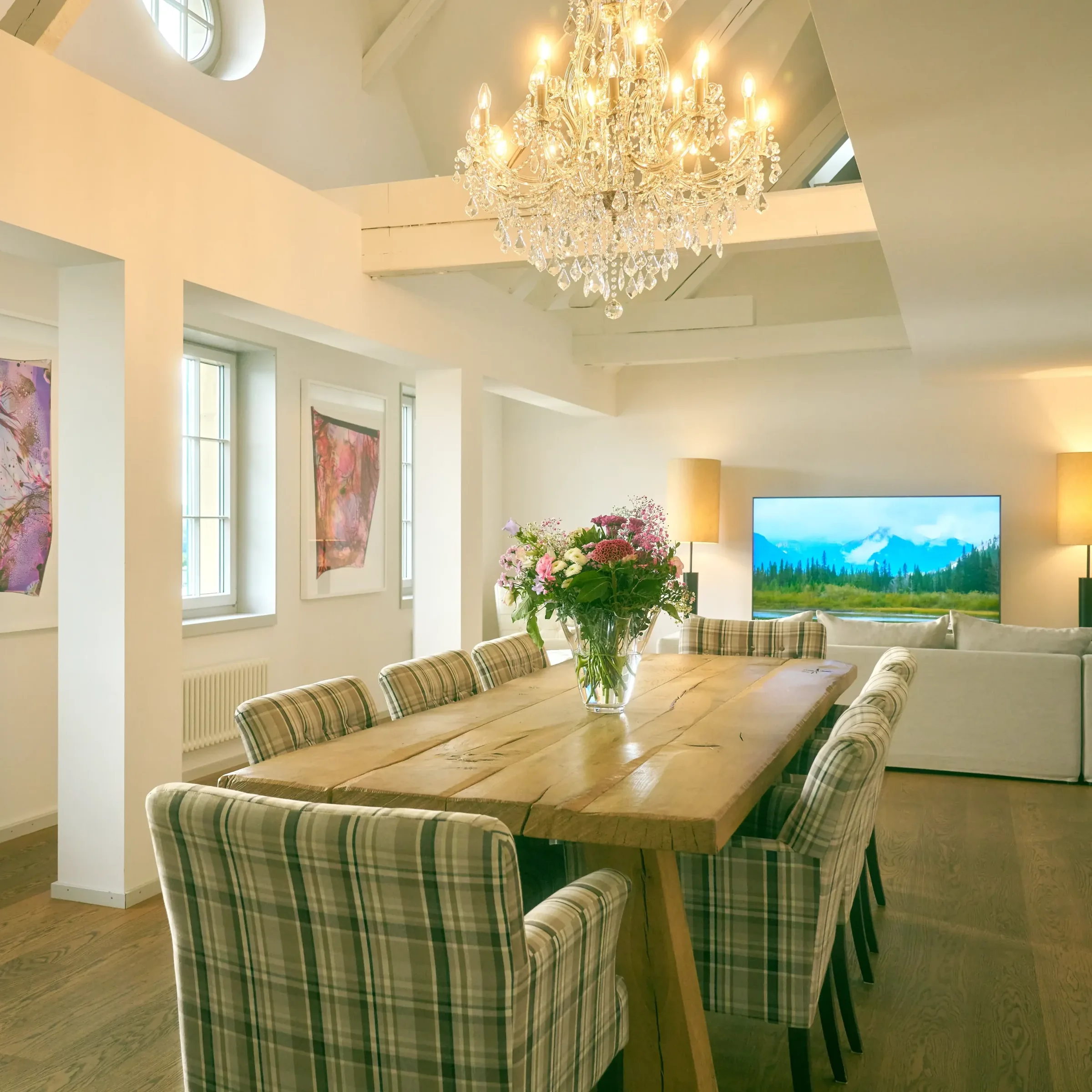 Open dining and living area with a long wooden table, checked chairs and a crystal chandelier