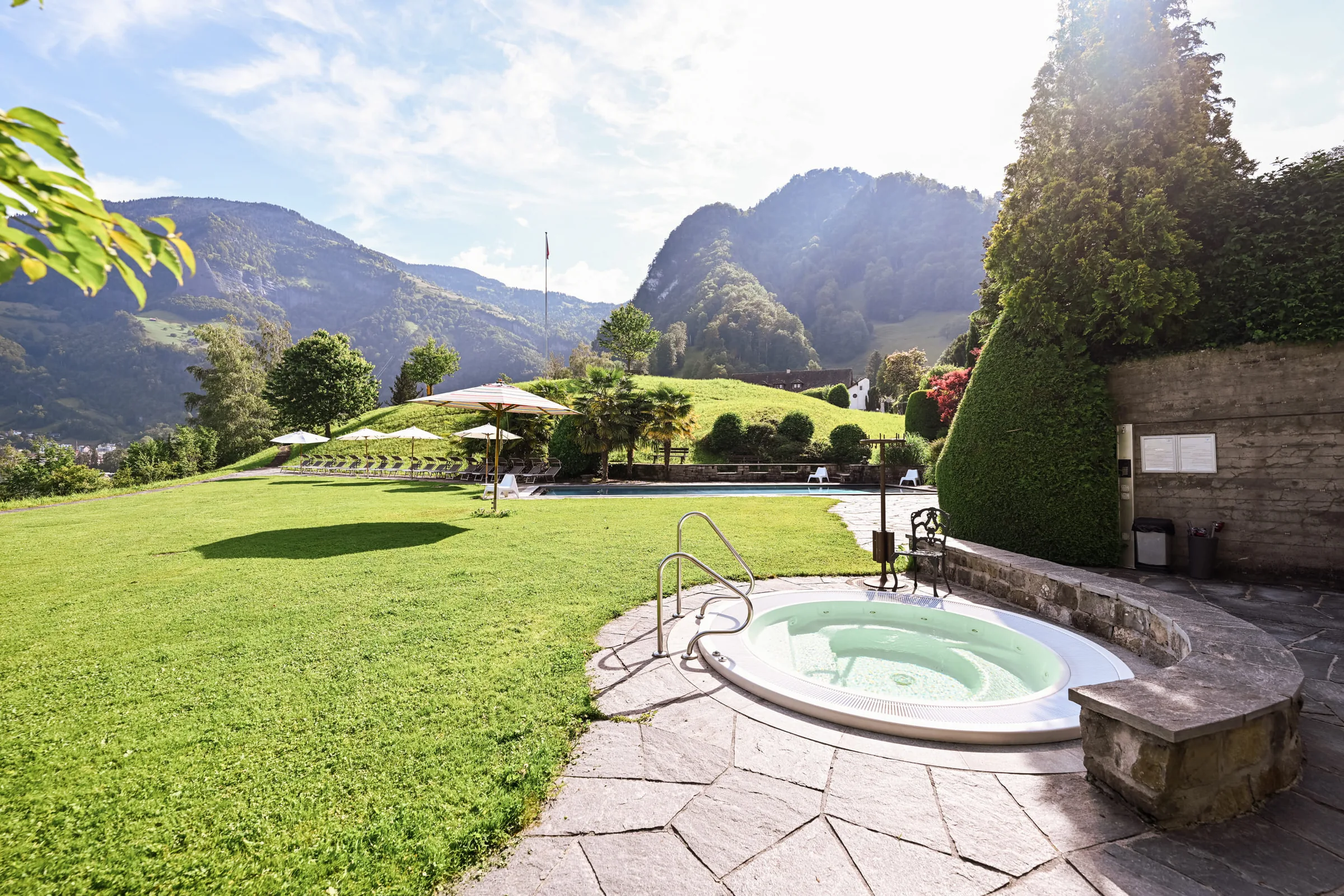 Round outdoor whirlpool set in stone paving on a sunny lawn, with parasols and mountains behind