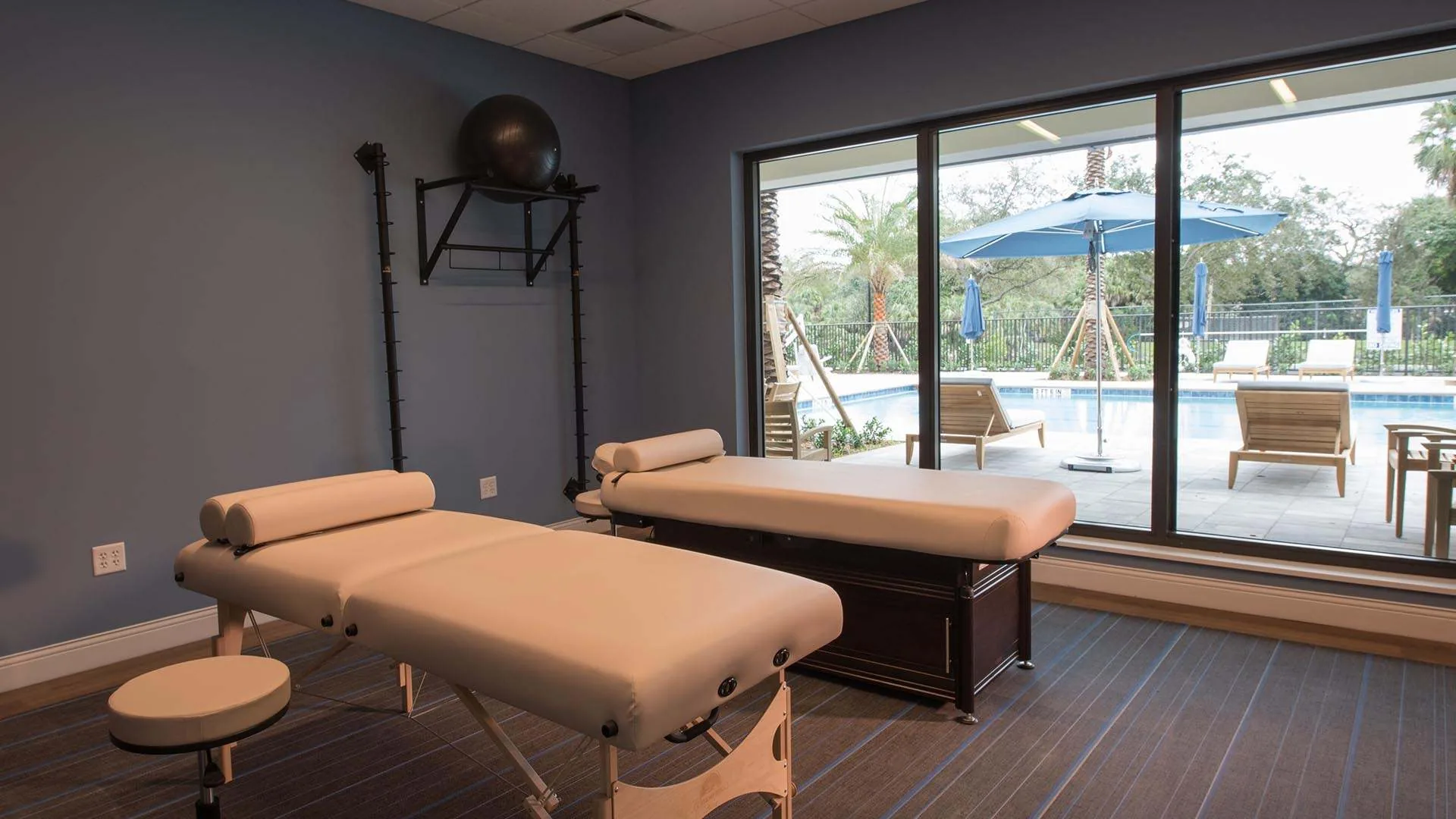 Massage room with two padded tables, grey walls and floor to ceiling windows facing the pool