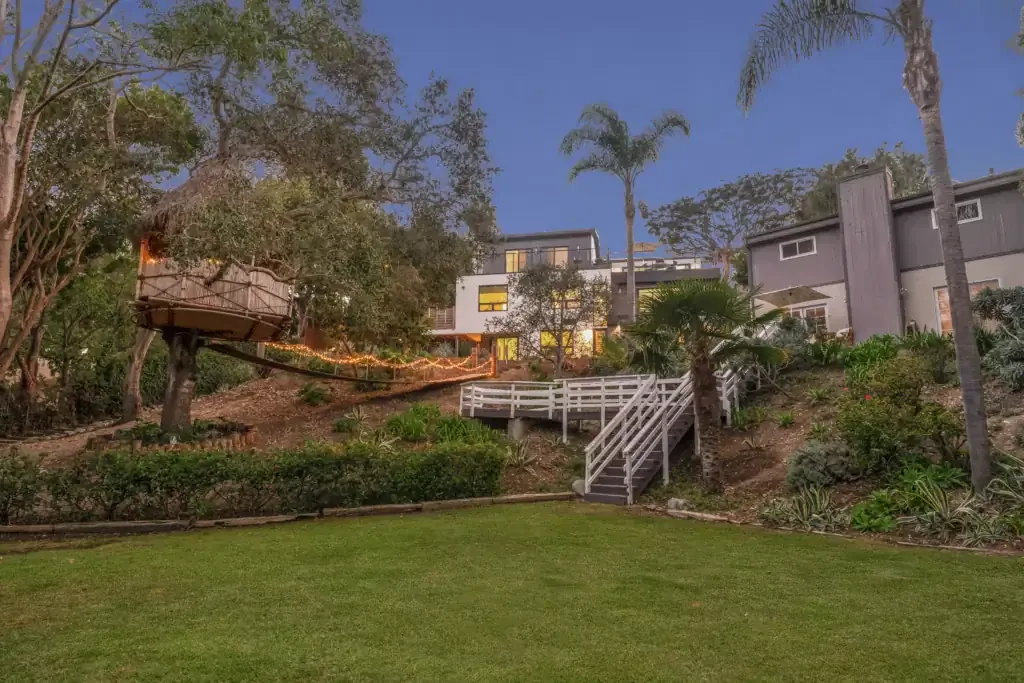 Lawn and wooded slope with a lit tree house below the Wildlife House residence at dusk in Malibu