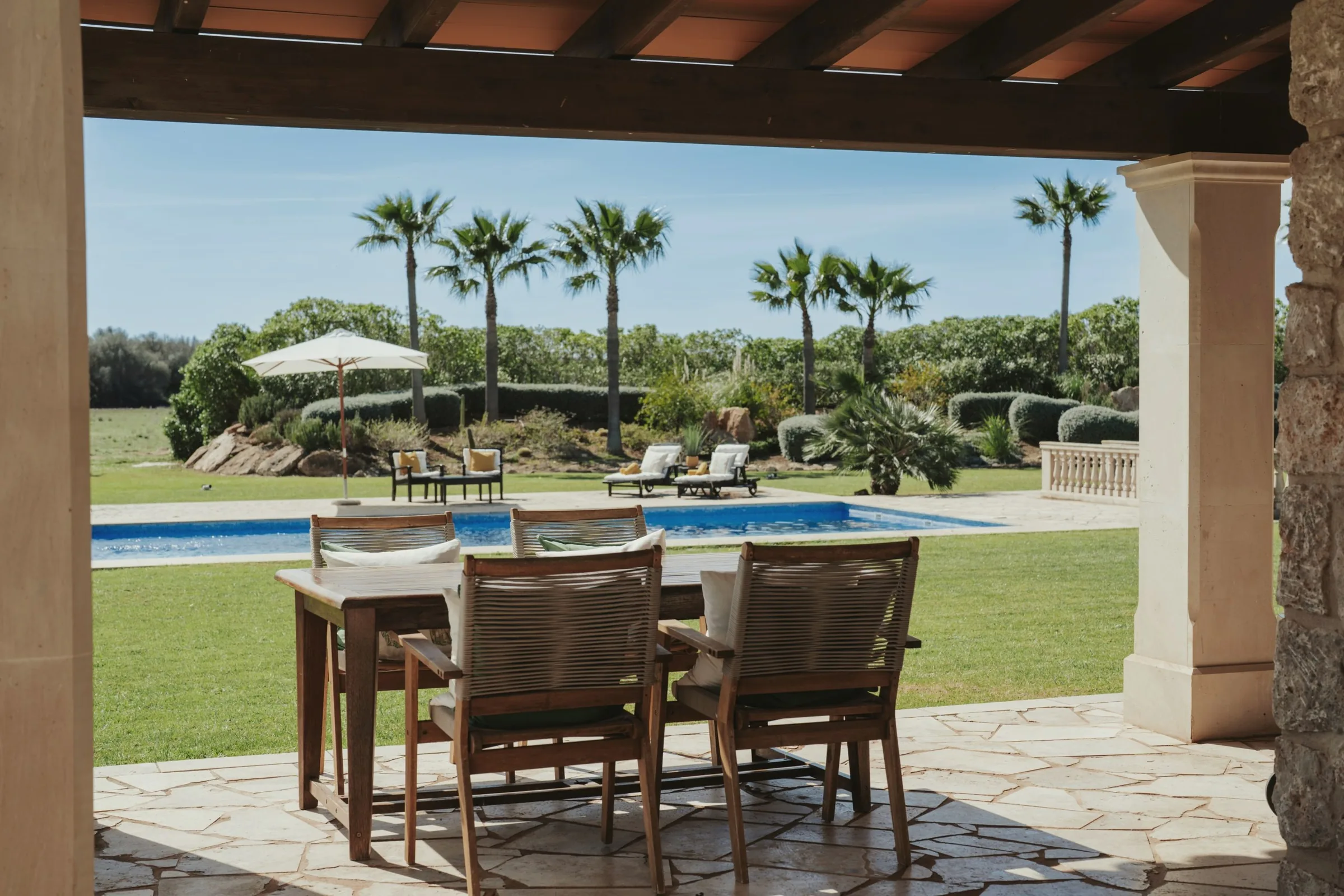 Covered stone terrace with a dining table looking onto a pool, lawn and palm trees