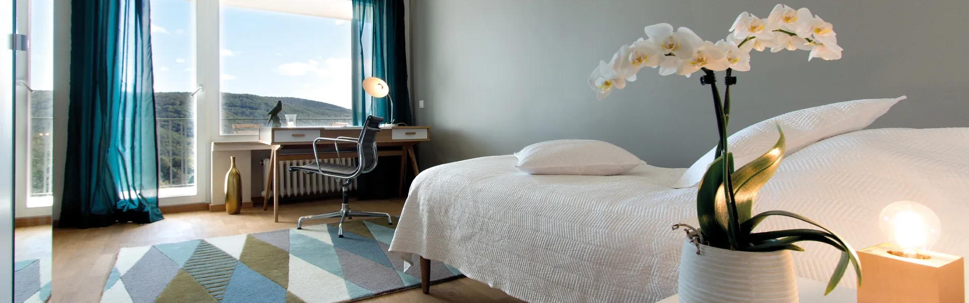 Single bedroom with a white bed, an orchid, teal curtains and a desk by a large window