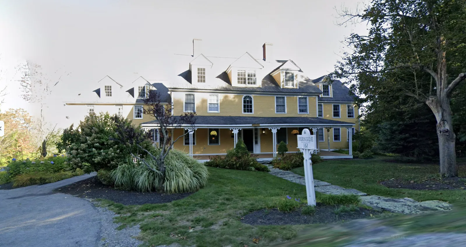 Yellow clapboard house with a wraparound porch and the Fernside 162 sign at McLean at Fernside, Princeton, Massachusetts