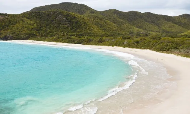 Curved white sand beach with turquoise water below green hills on the coast of Antigua