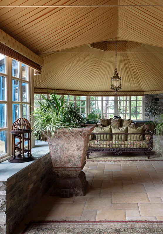 Conservatory with a stone floor, potted plants, a large urn and sofas under a pale timber ceiling