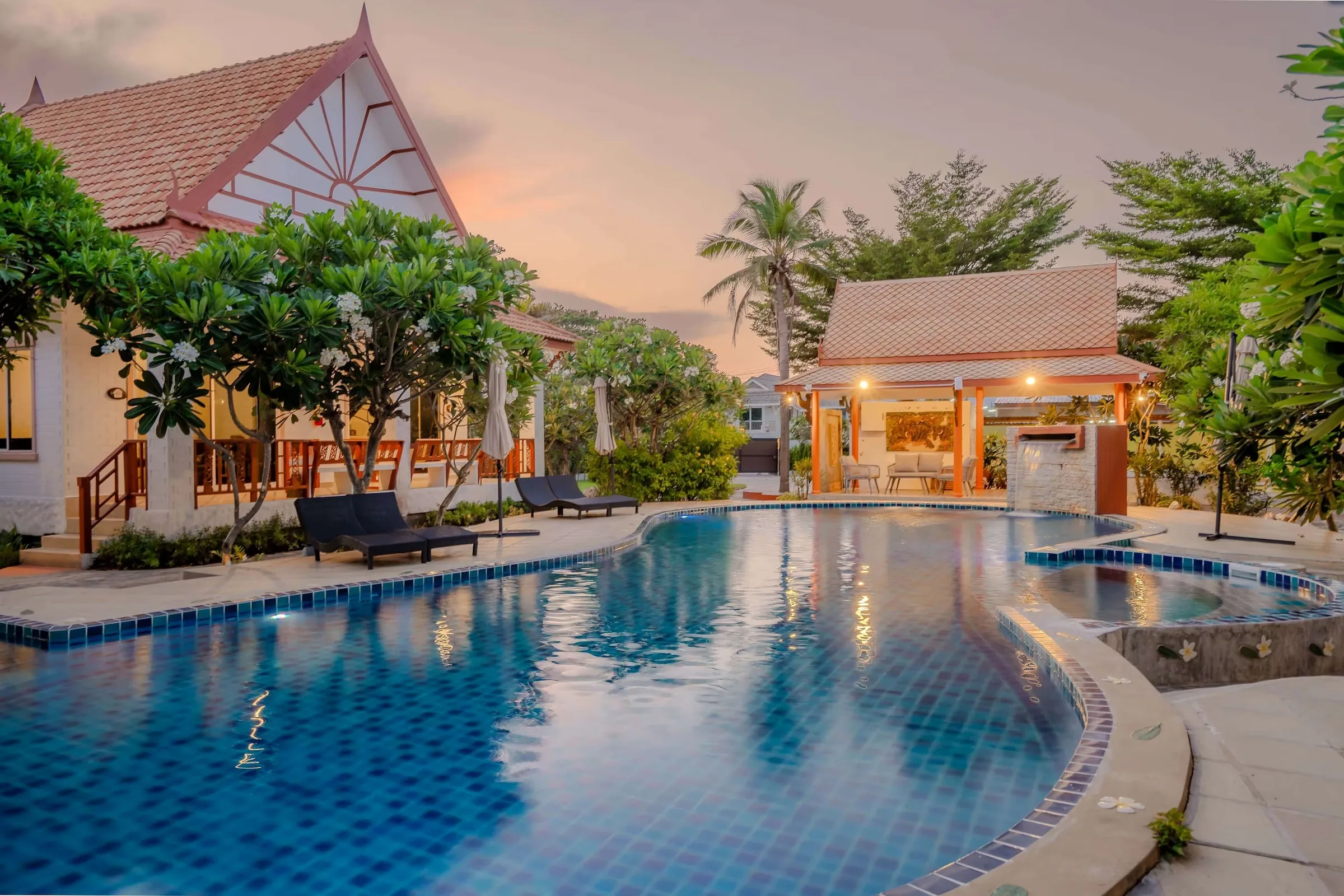 Curved swimming pool lined with sun loungers and palms, Thai-roofed bungalows behind under an evening sky