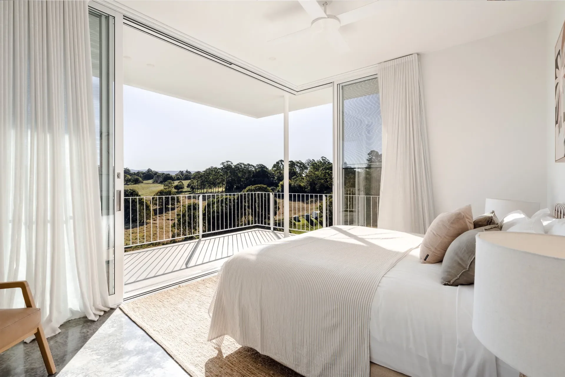 White bedroom with sheer curtains opening onto a balcony with views of green hinterland