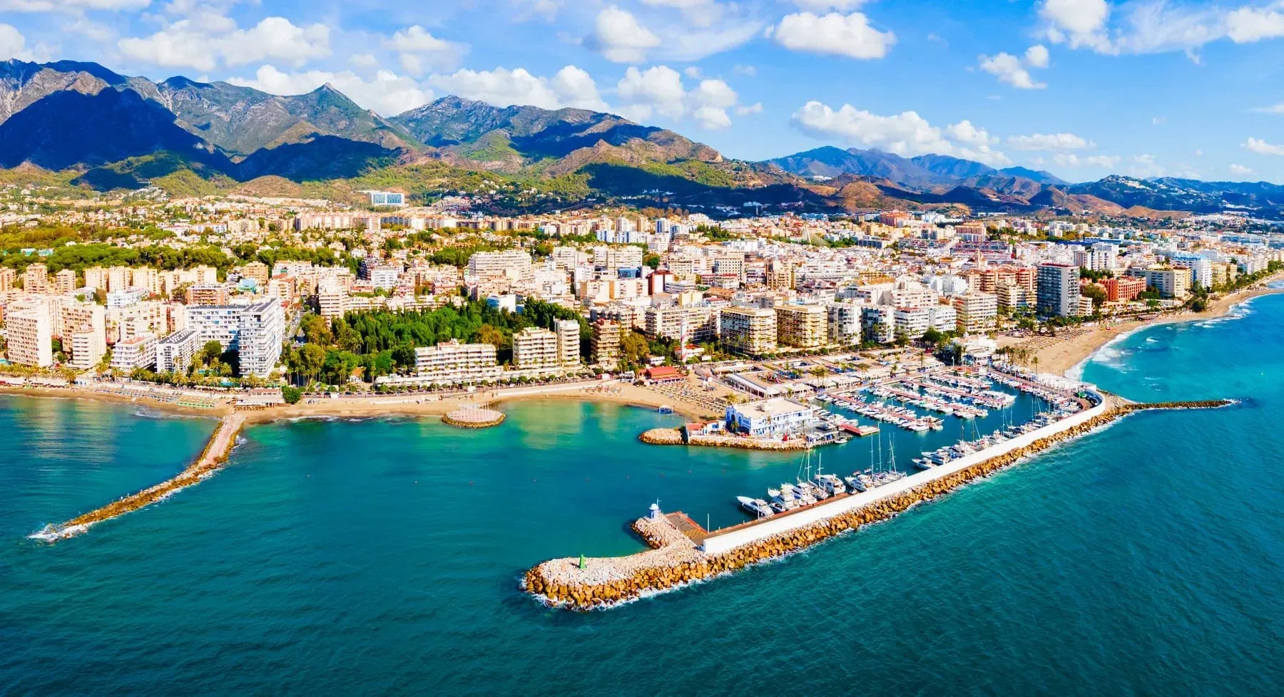Aerial view of a marina with yachts, beaches and apartment blocks along the coast below mountains