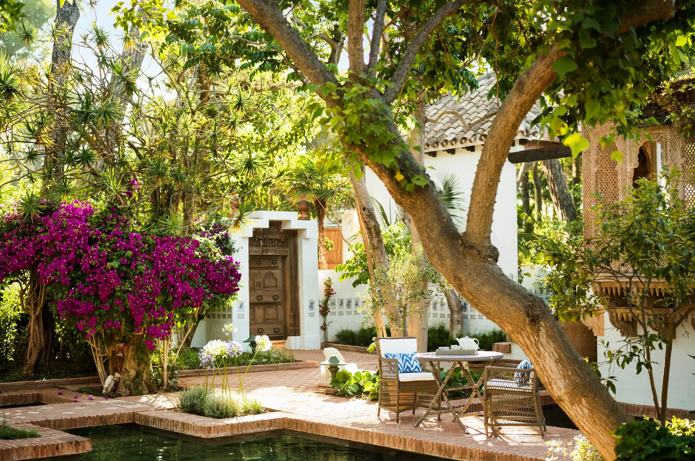 Brick garden patio with a table and chairs under old trees, purple bougainvillea and a white stucco house