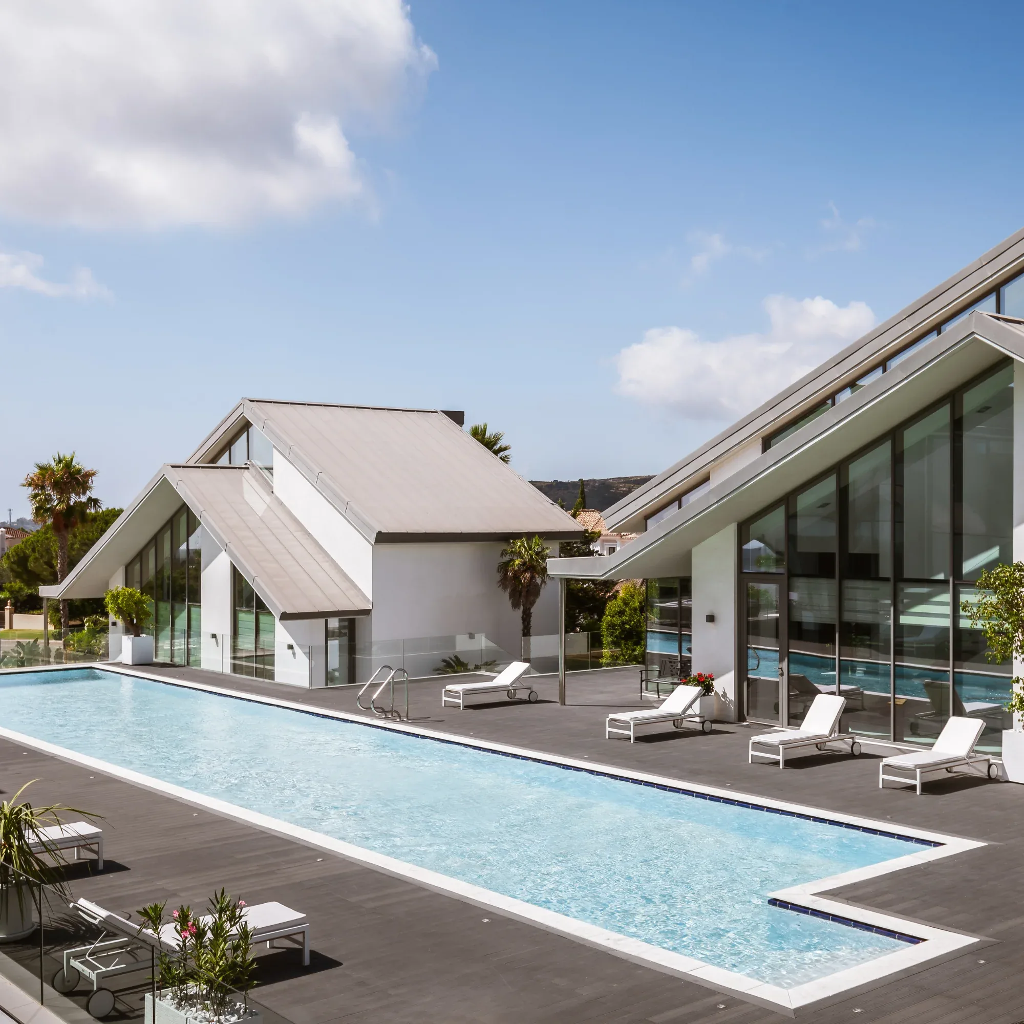 Long outdoor swimming pool with sun loungers on a wooden deck beside white pitched roof villas