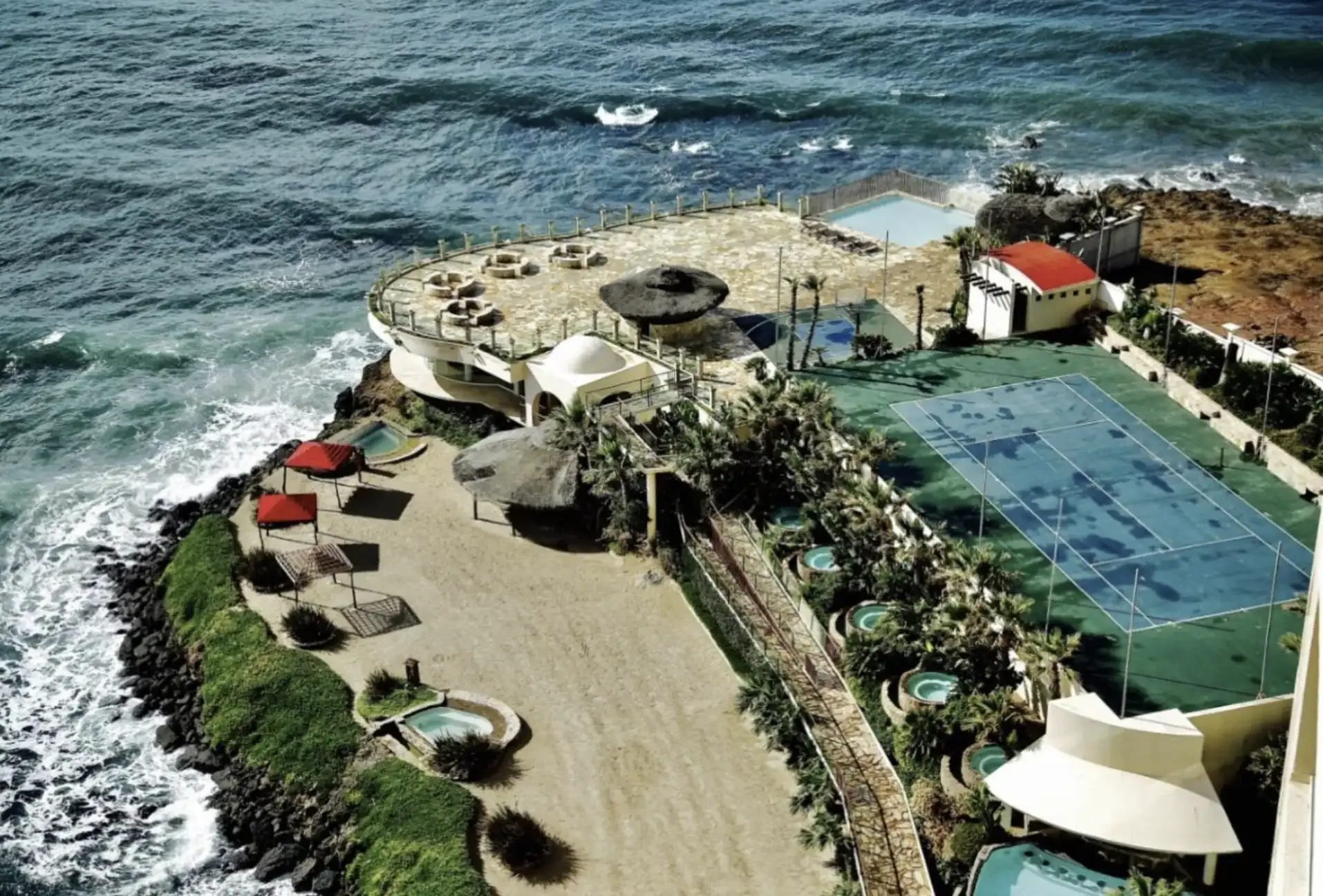 Aerial view of a cliffside property with a tennis court, terraces and pools above the Pacific surf