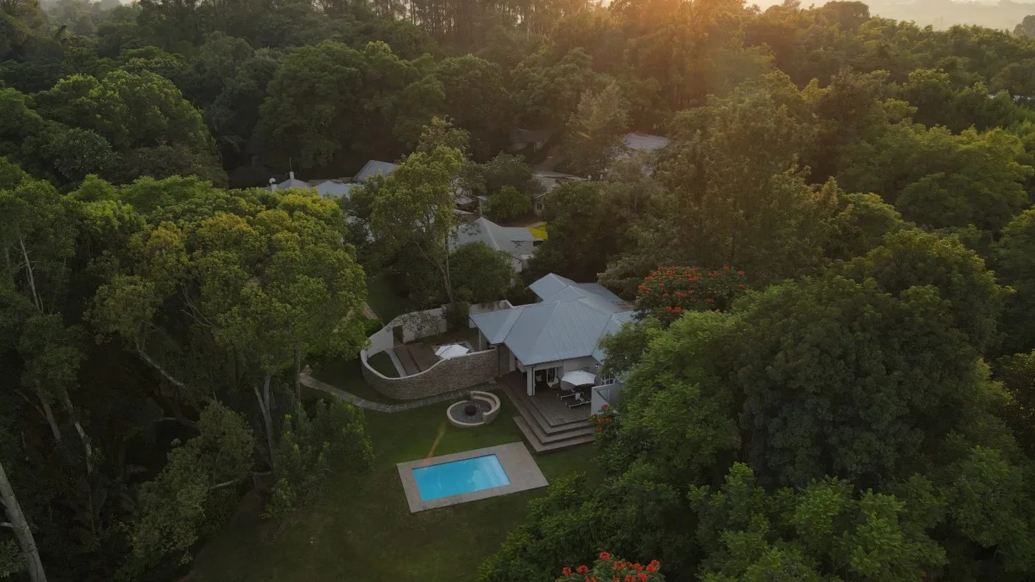 Aerial view of lodge buildings and a small pool among dense tall trees at sunrise