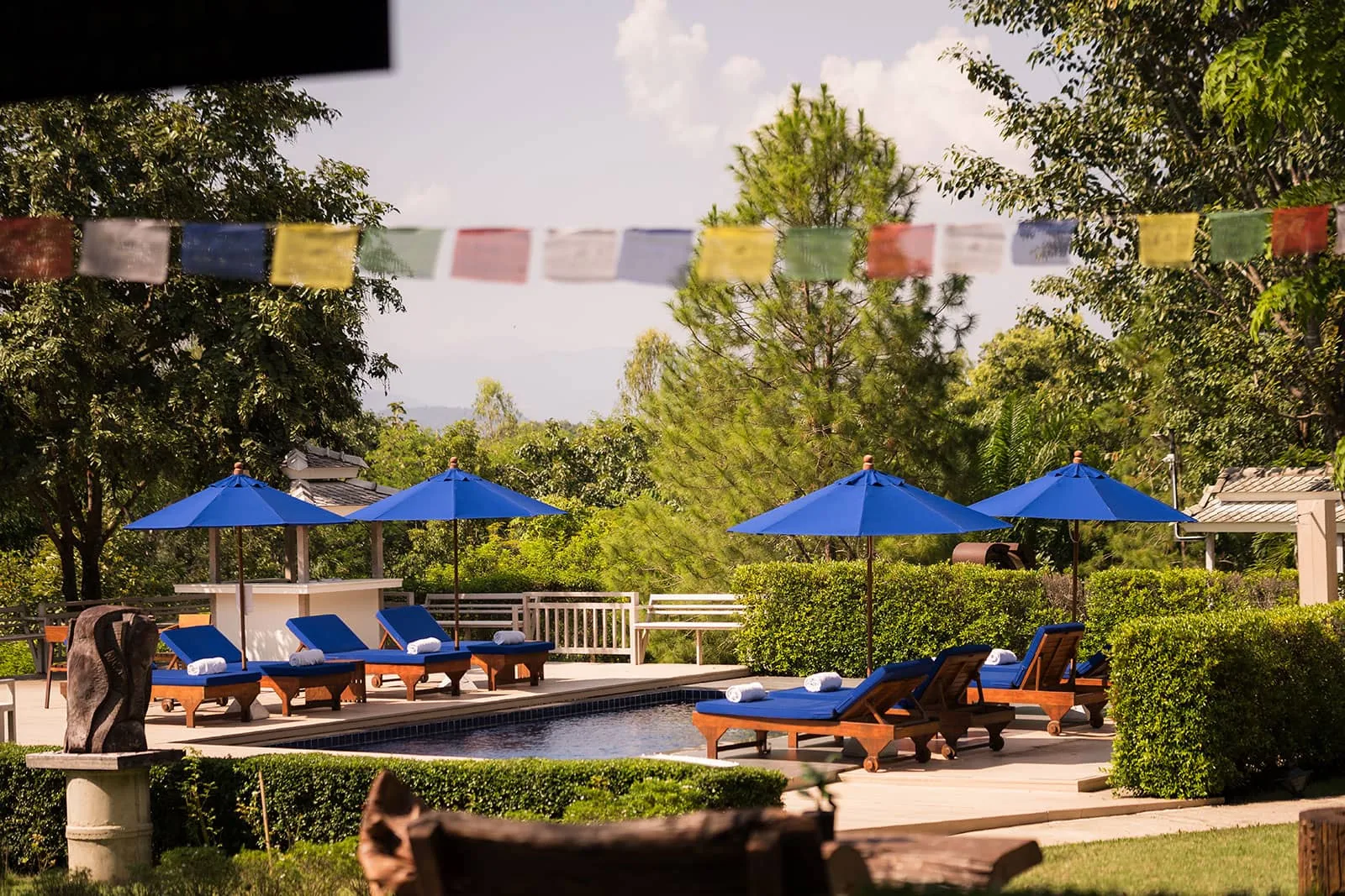 Outdoor swimming pool with blue parasols, sun loungers and prayer flags strung between garden trees