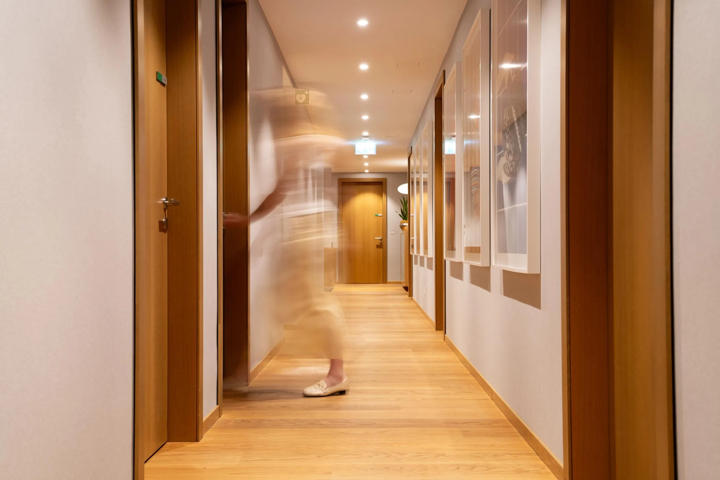 Long residence corridor with oak floors, wooden doors and framed pictures, a blurred figure walking