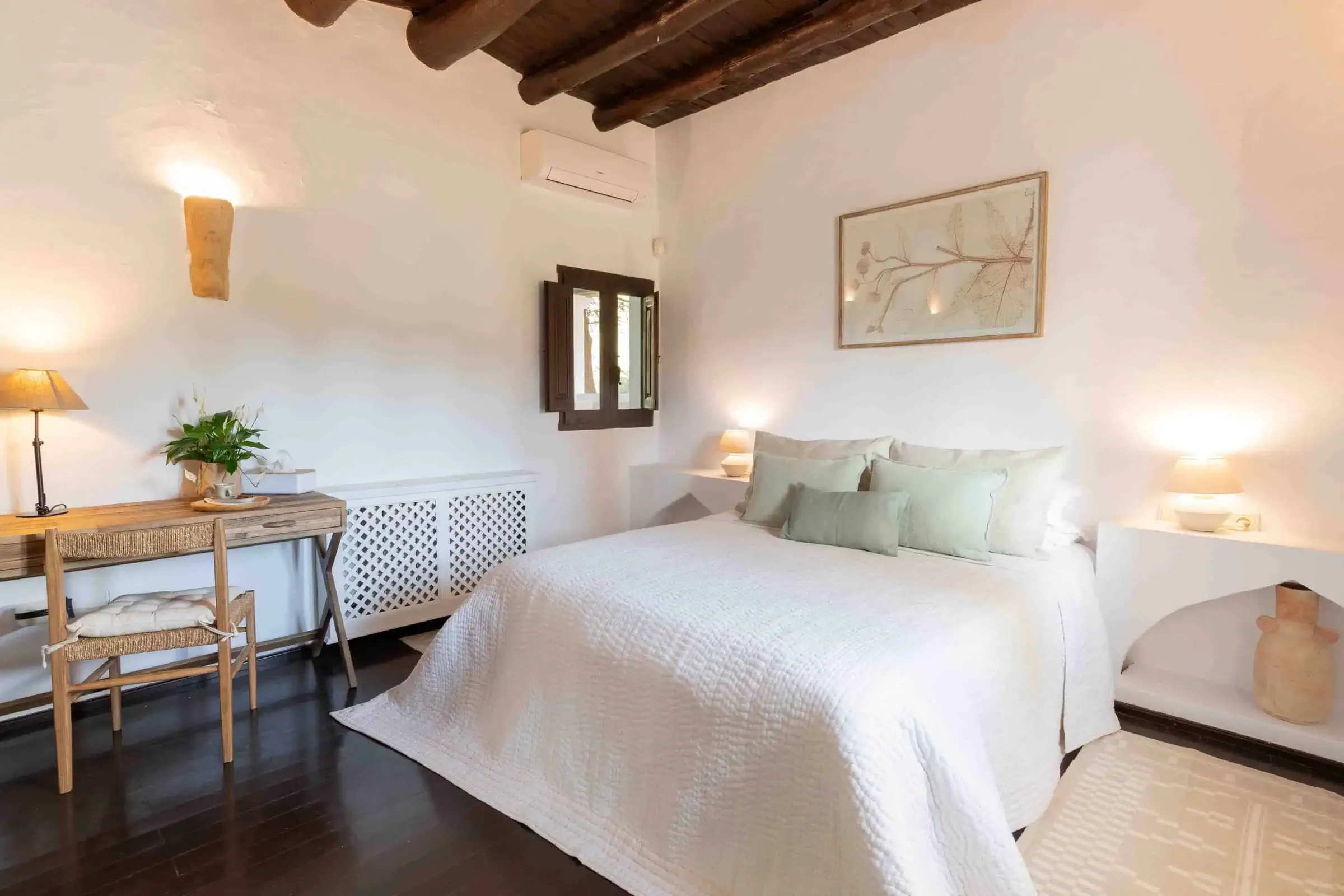 Bedroom with a white double bed, dark beamed ceiling, writing desk and wall lamps