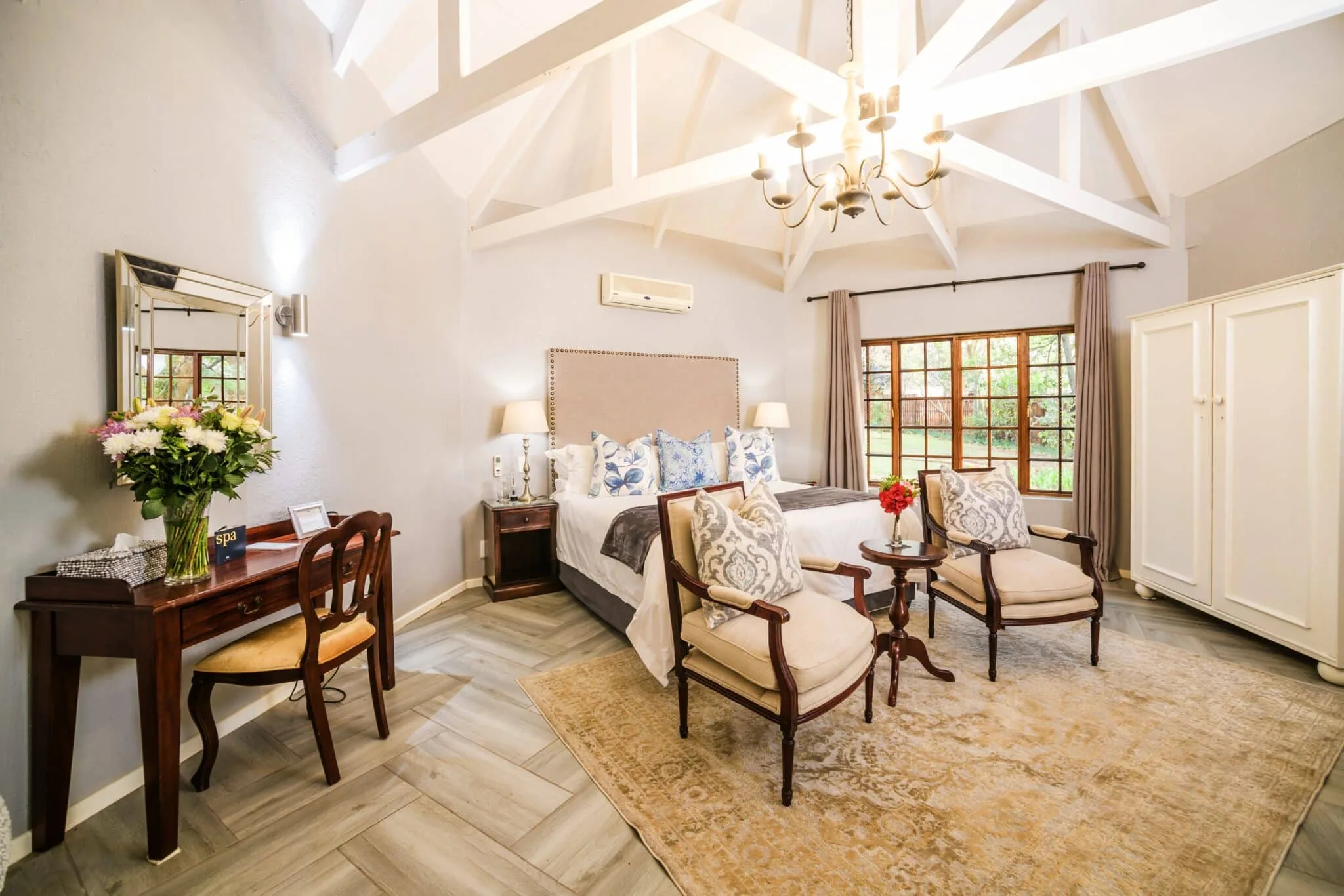 Suite with a vaulted white ceiling, chandelier, double bed, armchairs and a writing desk with flowers