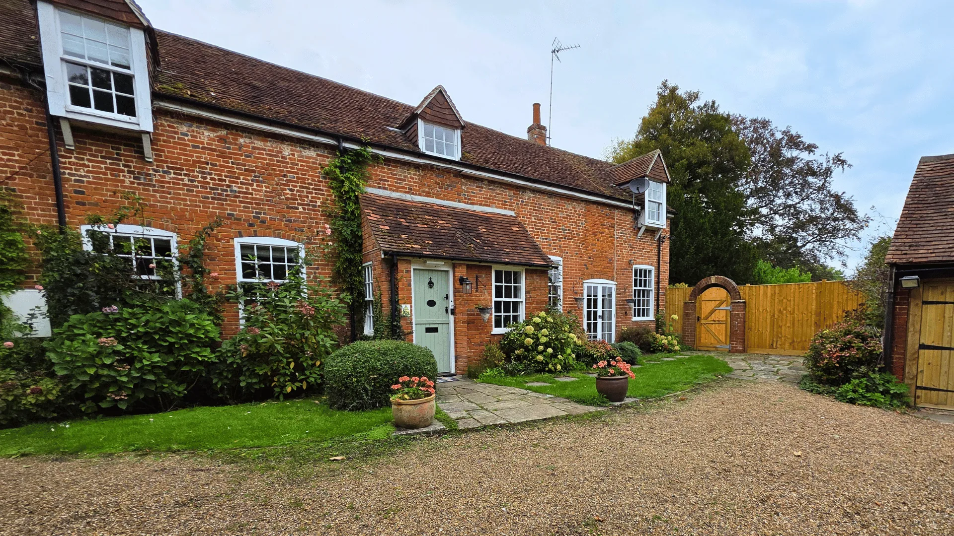 Red brick cottage with white windows, climbing plants and a gravel forecourt under a blue sky