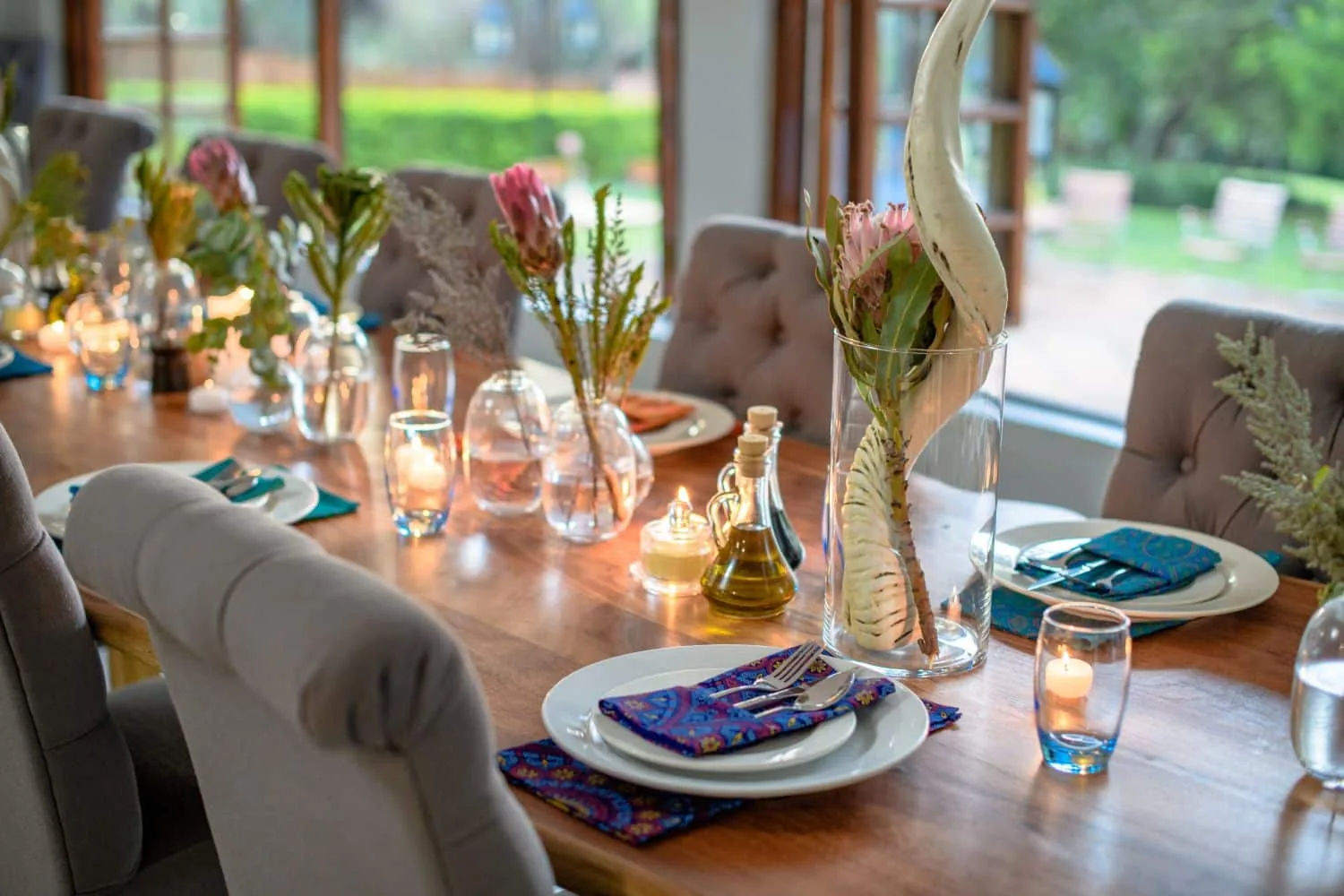 Long dining table laid with plates, glasses, candles and protea flowers beside garden windows