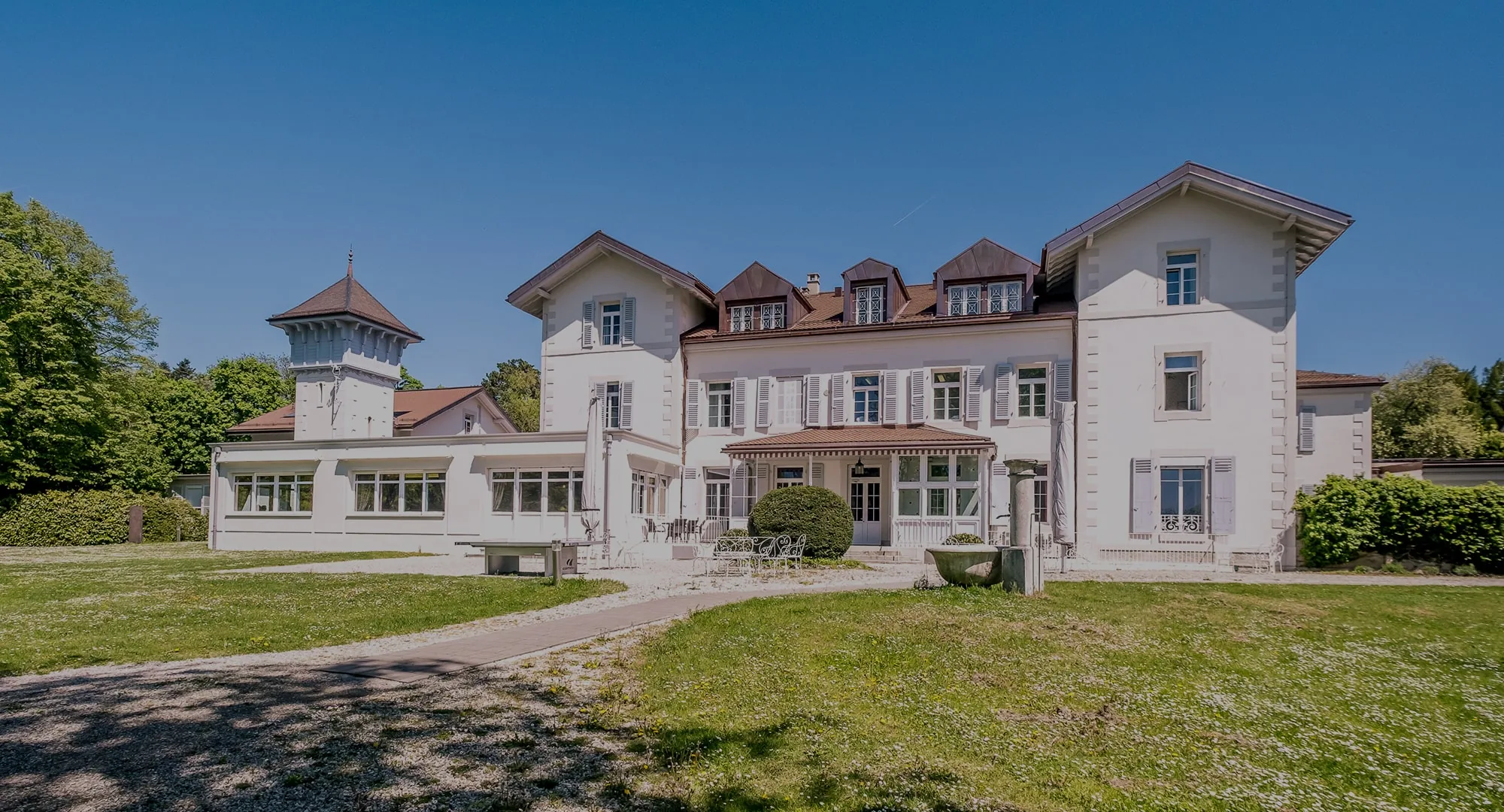 White historic villa with a turret and dormer windows behind a lawn and gravel path under blue sky in Nyon