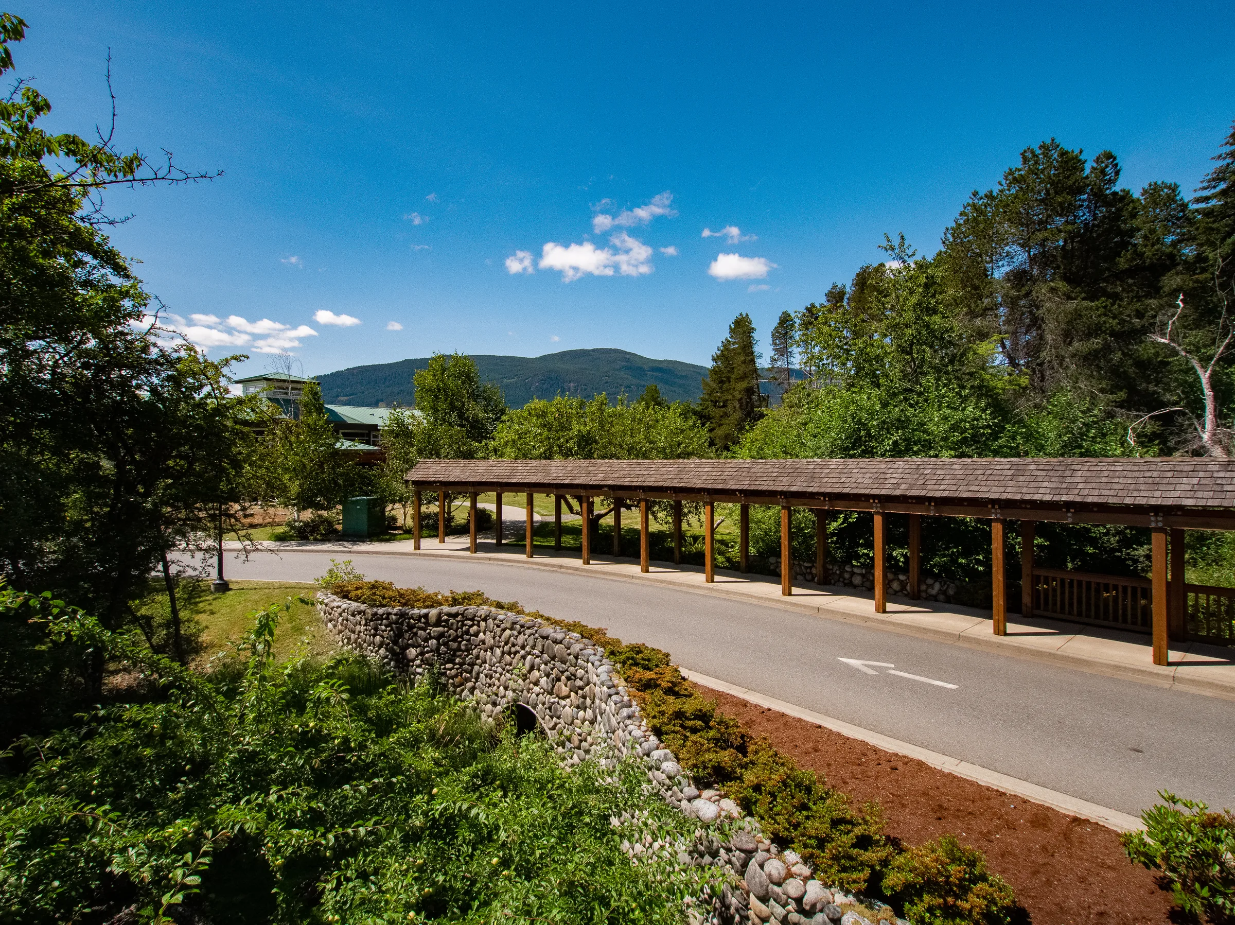 Long covered timber walkway beside a stone wall and road with forested hills and mountains behind