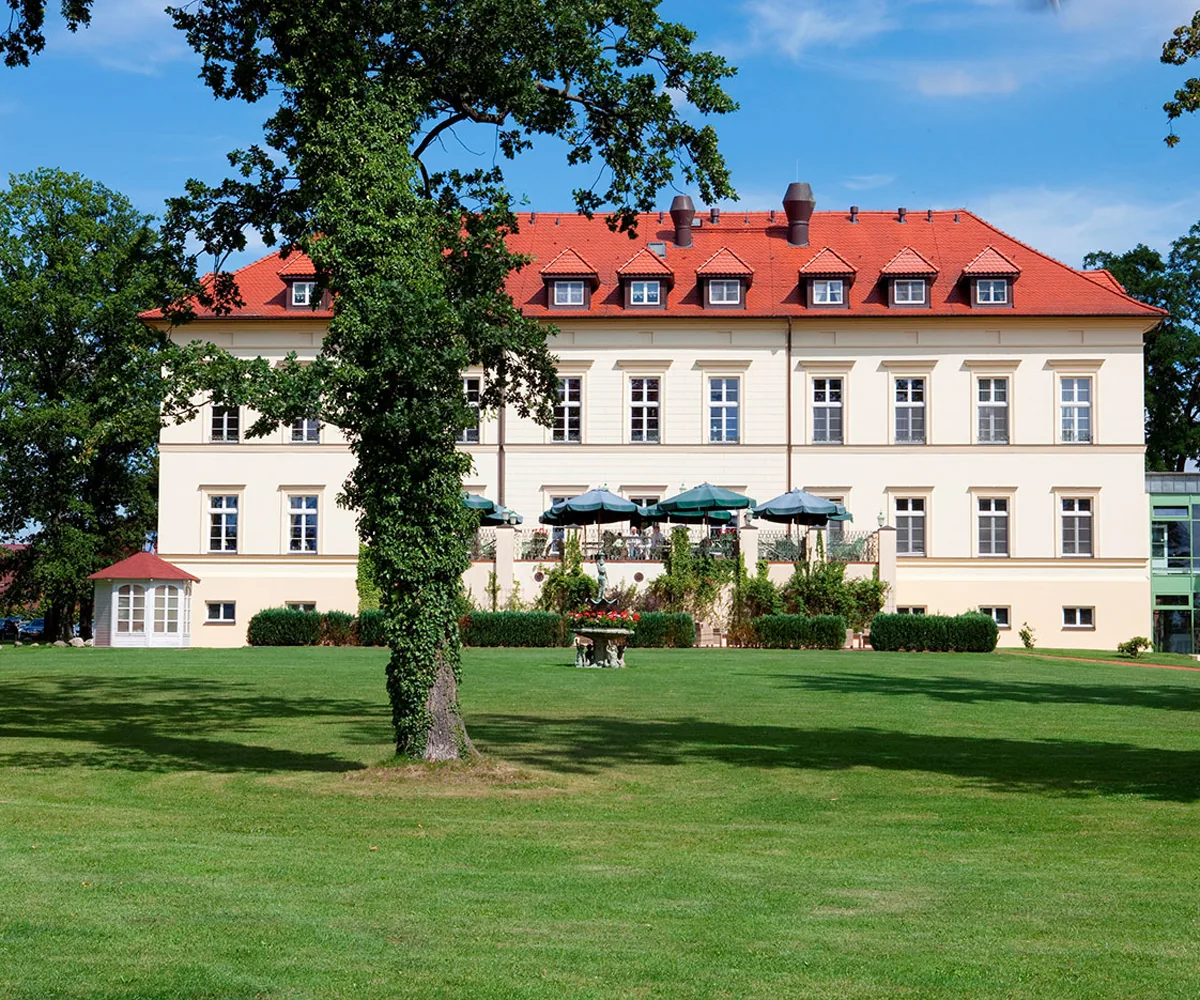 White manor house with a red roof and a terrace behind a lawn and old trees in Mecklenburgische Schweiz