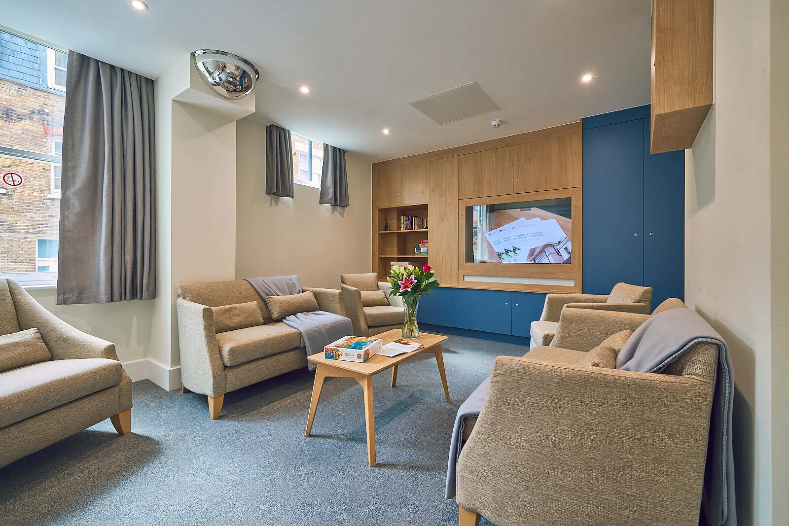 Ward lounge with upholstered armchairs, a wooden coffee table, bookshelves and a wall-mounted screen