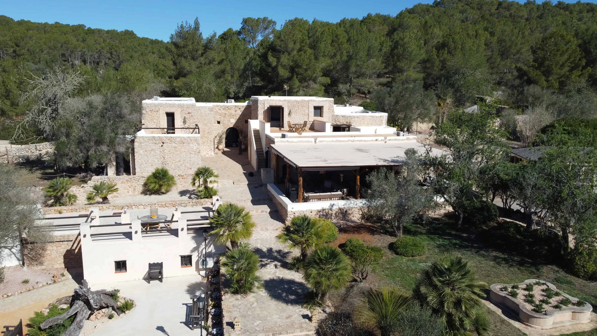Aerial view of low white terraced buildings, gardens and a drive set in pine woodland on Ibiza