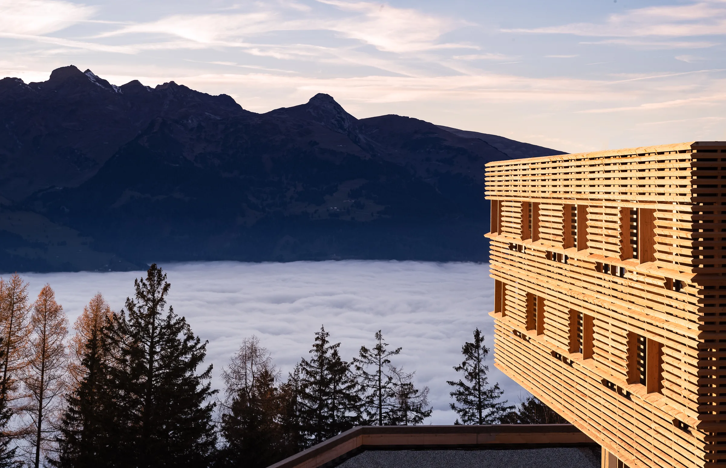 Timber-clad clinic facade at dusk above a sea of fog in the Rhine valley with mountain ridges behind