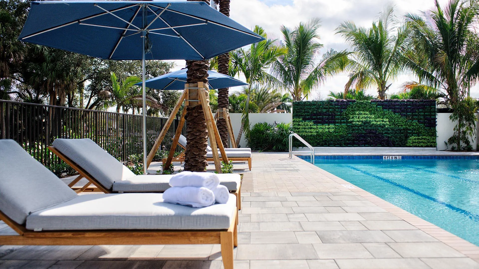 Pool deck with cushioned loungers, folded towels, blue umbrellas, palms and a green living wall