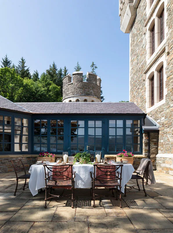 Courtyard dining table with red chairs beside a stone castle wall, a glass pavilion and a turret