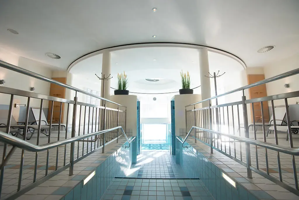 Indoor swimming pool with steel handrails and steps leading down into the water under a curved white ceiling