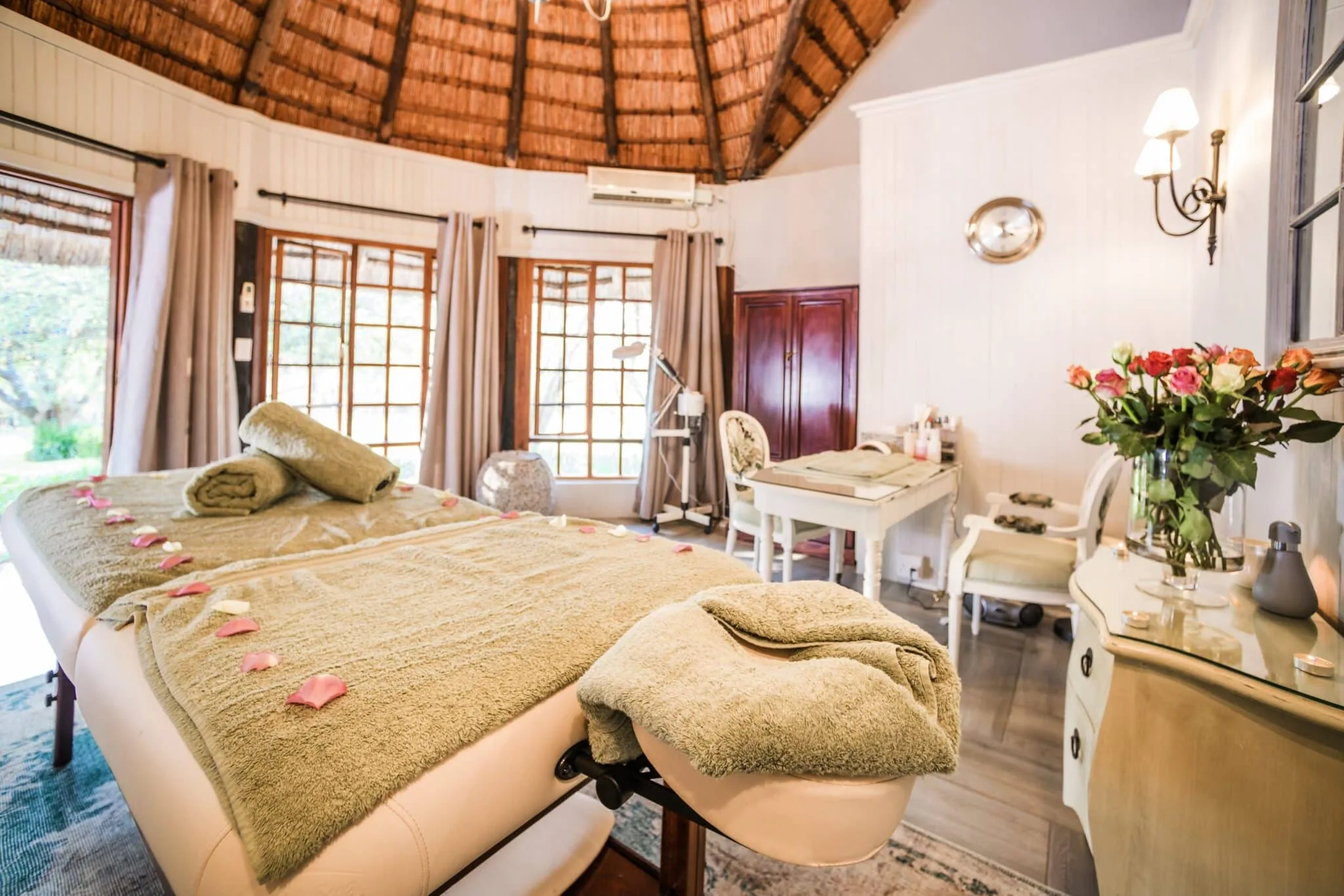 Spa treatment room under a thatched roof with a massage bed, folded towels, petals and roses