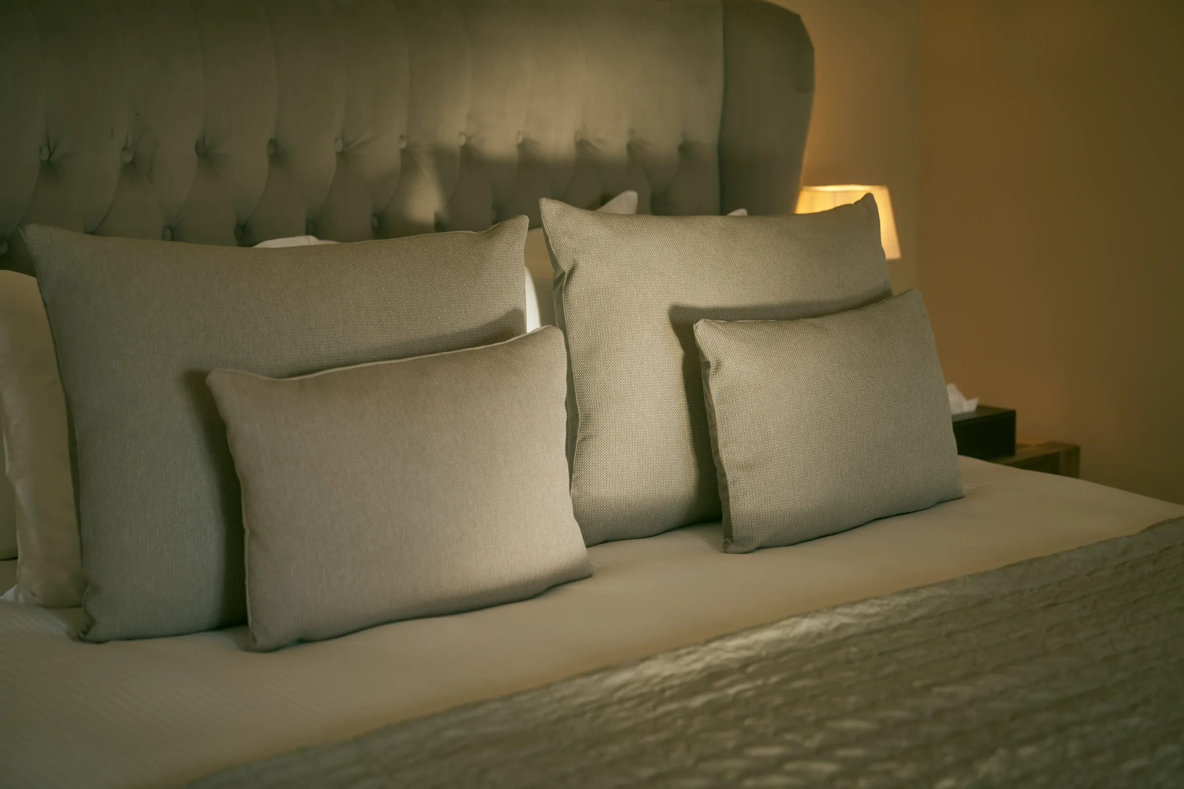 Double bed with a tall upholstered headboard and grey cushions lit by a warm bedside lamp
