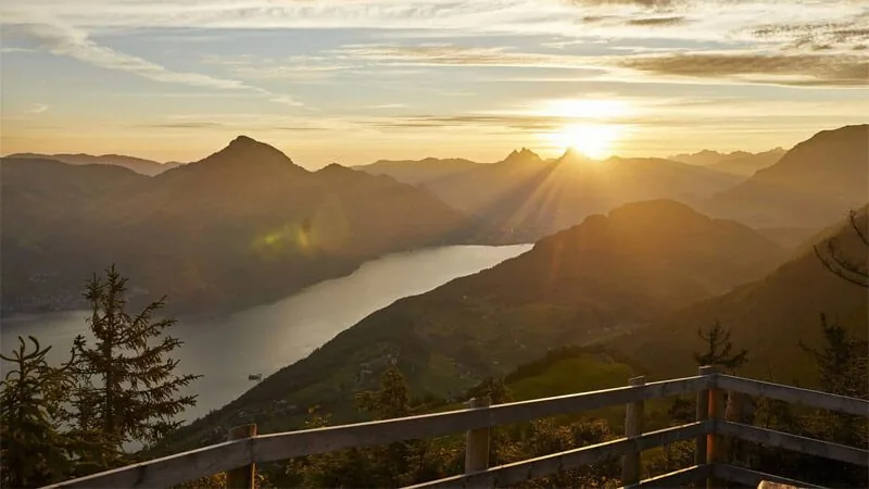 Sun setting over mountain ridges and an arm of Lake Lucerne, seen from a wooden balcony railing