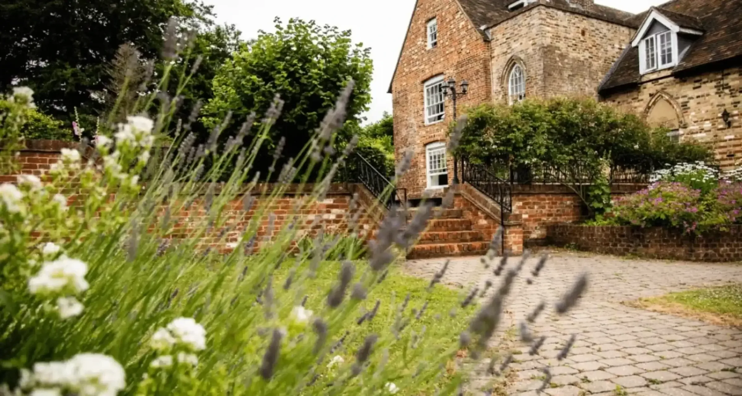Brick and stone farmhouse behind a paved path lined with flowering lavender and white daisies