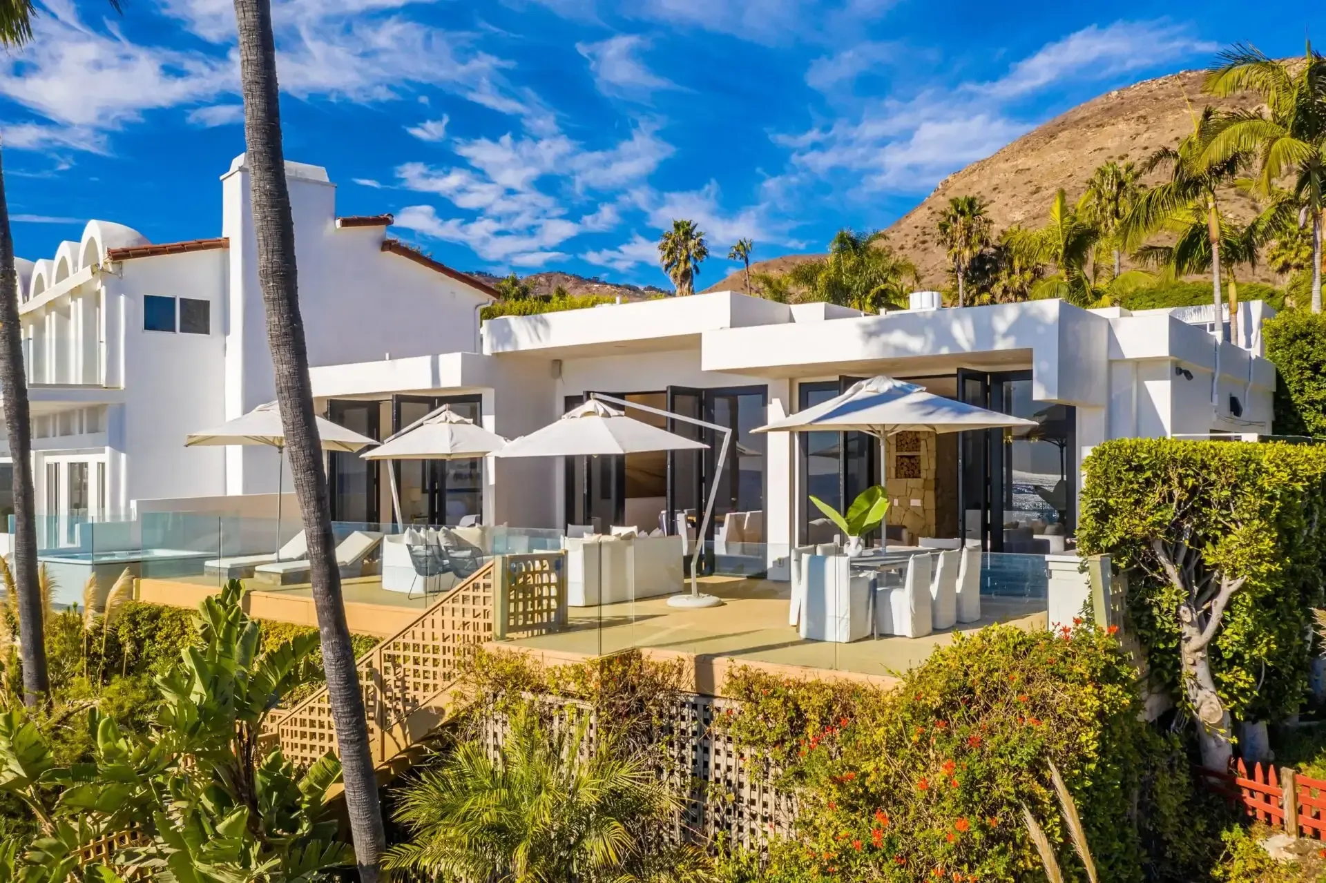 White modern house with glass railed terraces, umbrellas and palms on a green hillside under blue sky