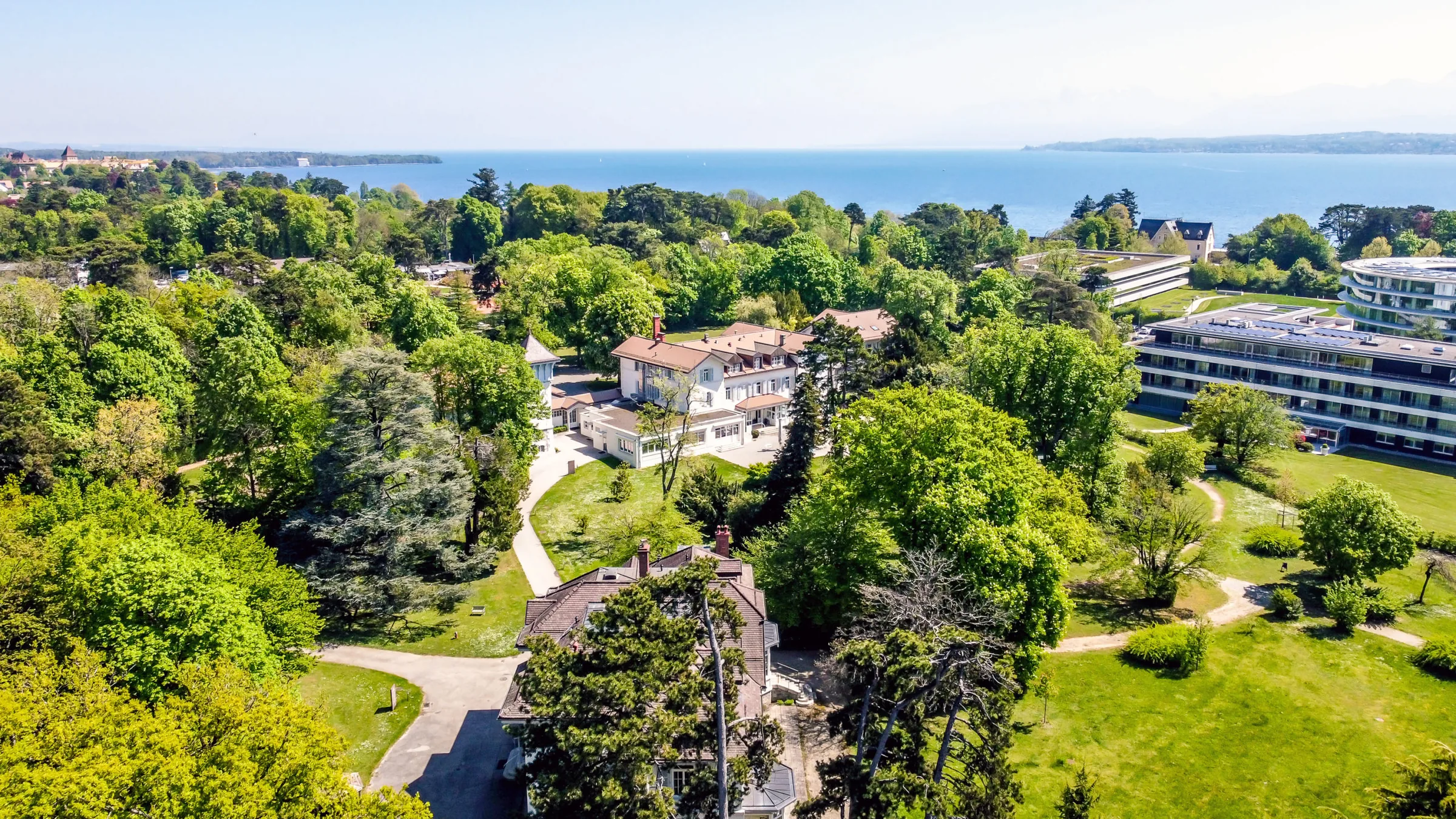 Aerial view of the clinic buildings set in a wooded park with Lake Geneva and the far shore beyond