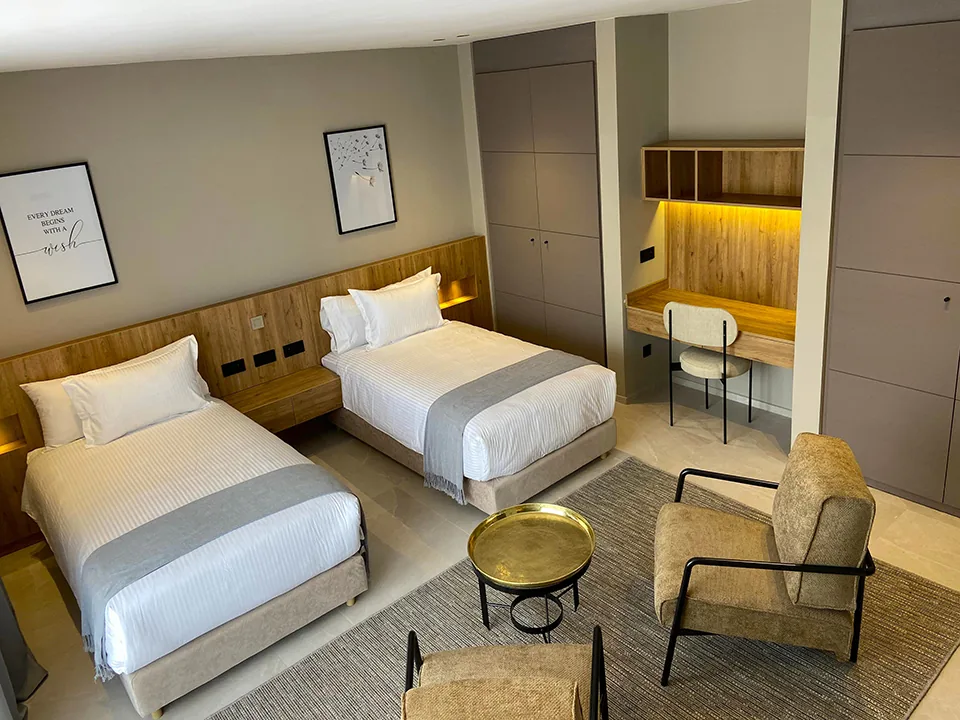Bedroom with two single beds in white linen, a desk, framed prints and two armchairs